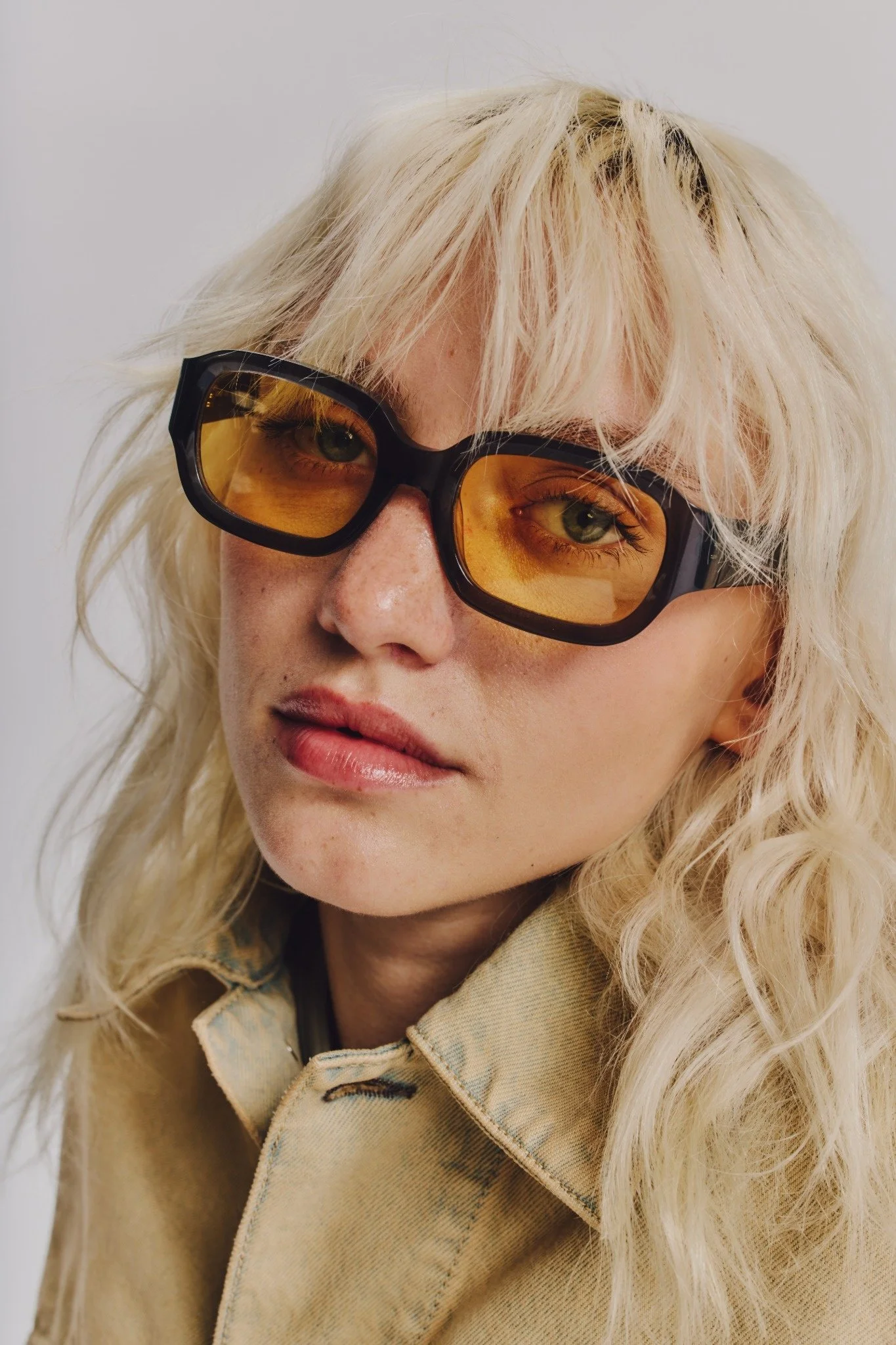 Close-up of a person with long, wavy blonde hair wearing large, yellow-tinted sunglasses and a beige jacket, against a plain light background.
