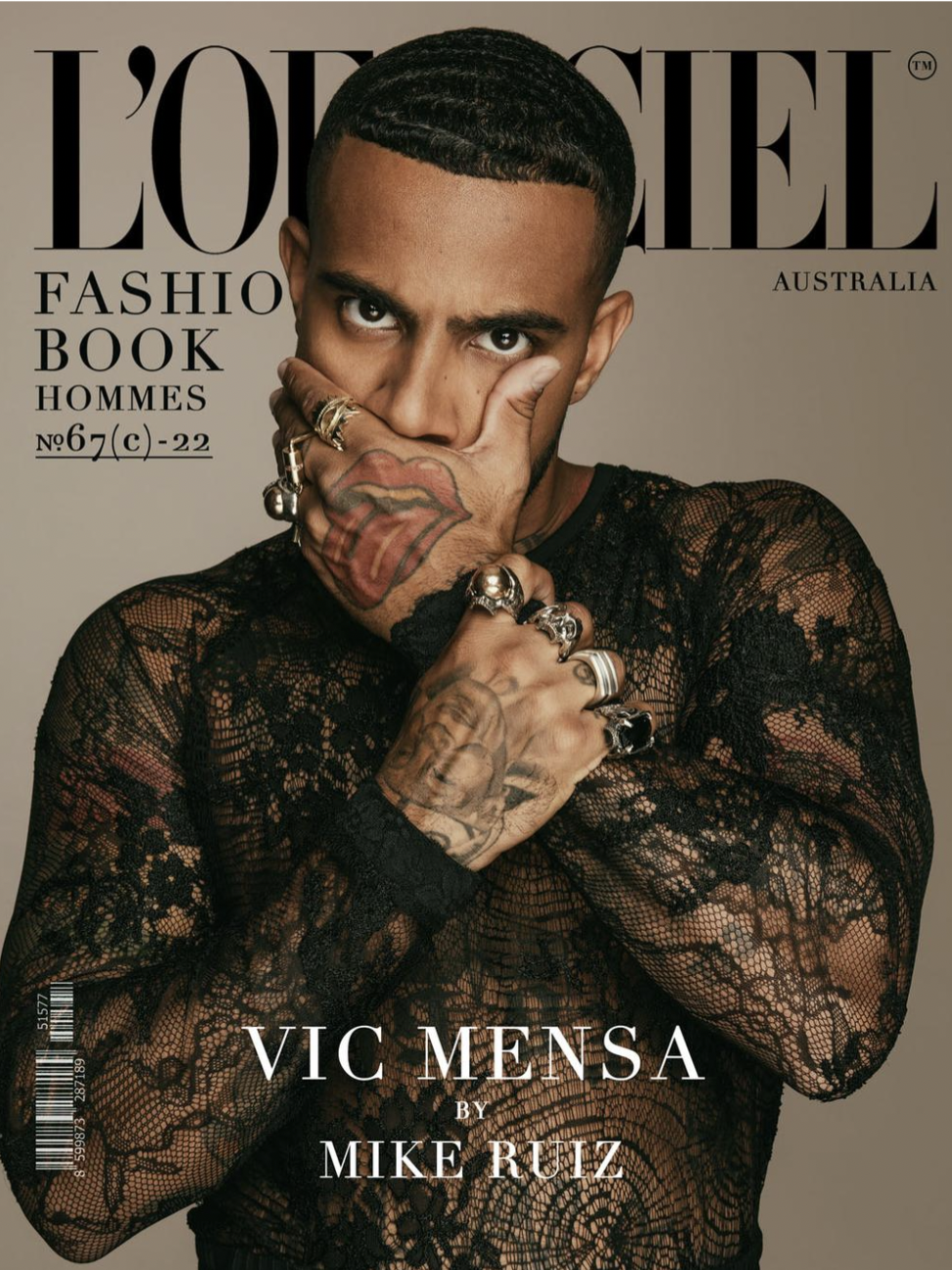 Close-up of a man with tattoos and multiple rings on fingers, wearing a black lace top, holding one hand over his mouth, on the cover of L'Officiel Australia magazine, featuring Vic Mensa by Mike Ruiz.