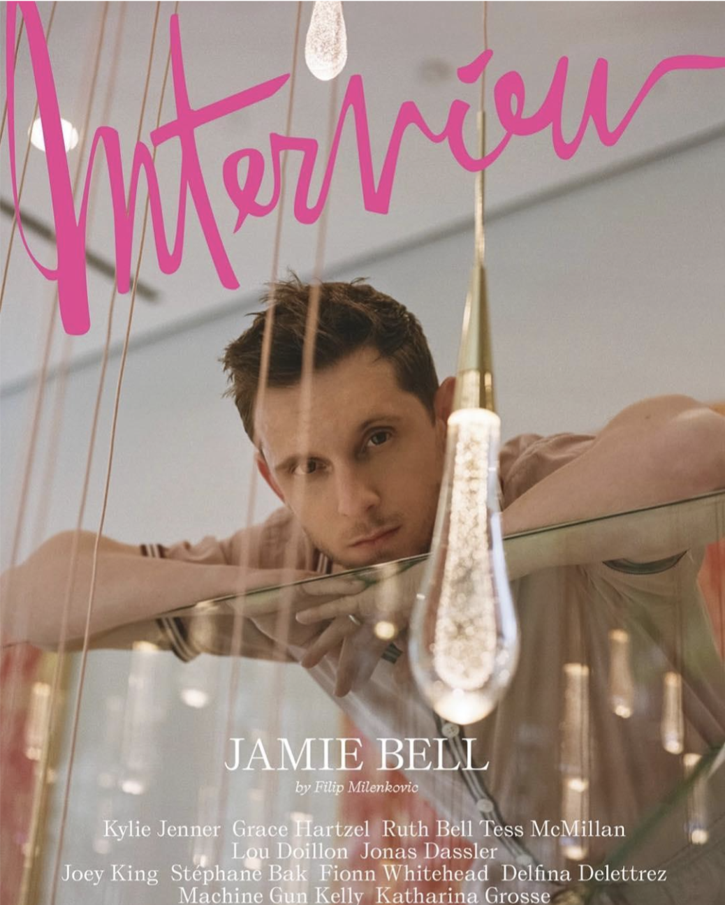 Magazine cover featuring a young man with short hair looking over a reflective surface with hanging lights and pink handwritten text that says 'interview.' The cover text includes 'JAMIE BELL' and lists of other names.