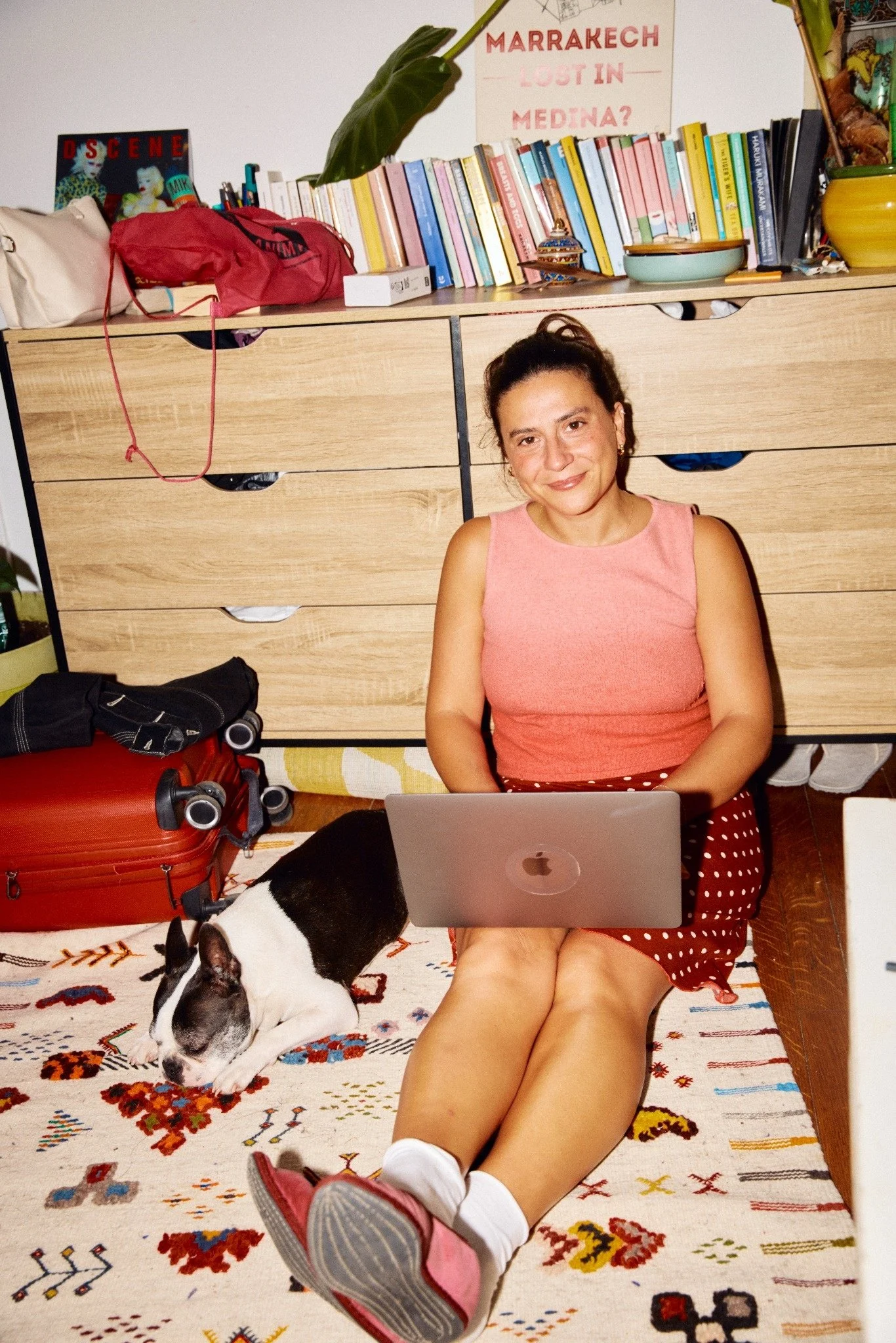 A woman with dark hair tied back, wearing a pink sleeveless top and red polka dot shorts, sitting on a colorful rug with her legs extended, using a silver laptop on her lap. A black and white dog is lying beside her. In the background, a wooden dresser with a lush green plant, a stack of books, and decorative items is visible. A poster with red text reads 'Marrakech Lost in Medina?' and another magazine titled 'Descene' is also on the dresser. There is a red suitcase and a black item, possibly a bag or clothing, on the floor. The woman is smiling at the camera.