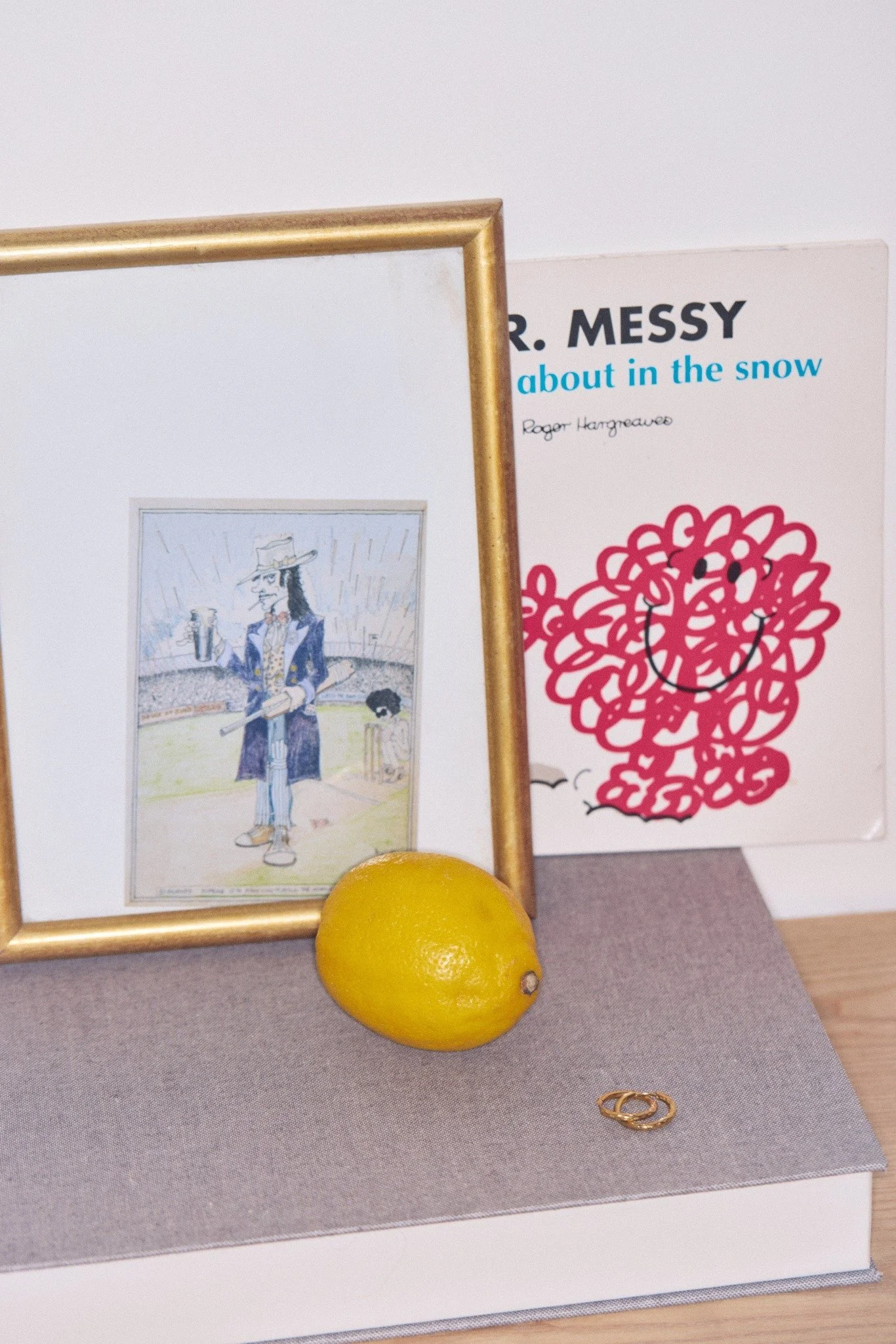 A yellow lemon, a framed mirror, and two gold rings on a gray display stand. In the background, there are posters, one with a cartoon drawing of a person in a blue coat and hat holding a drink, and another with abstract red scribbles and black eyes.