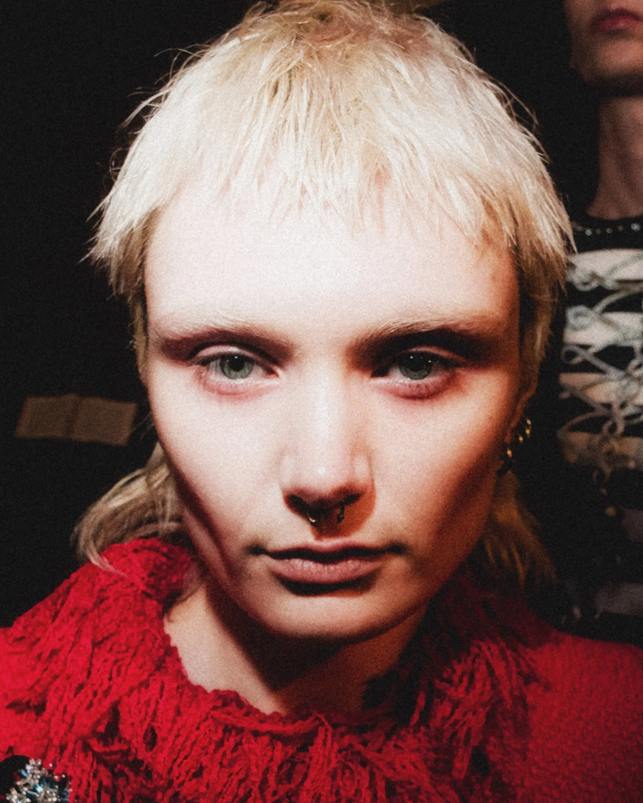Close-up of a person with platinum blonde hair, green eyes, and septum piercing, wearing a red textured sweater, against a dark background.