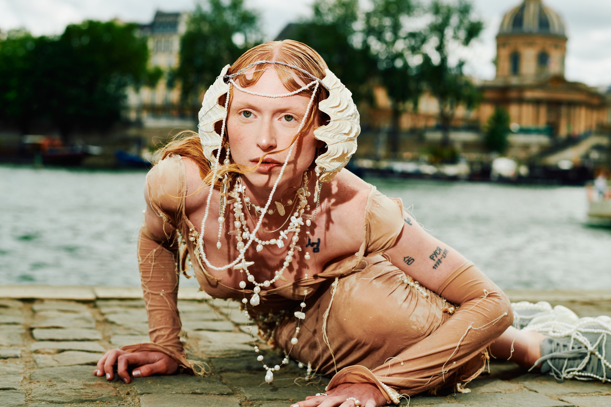 A person with long red hair in a beige outfit, adorned with multiple strands of pearls and jewelry, crawling on an outdoor stone surface near water with boats, trees, and historical buildings in the background.