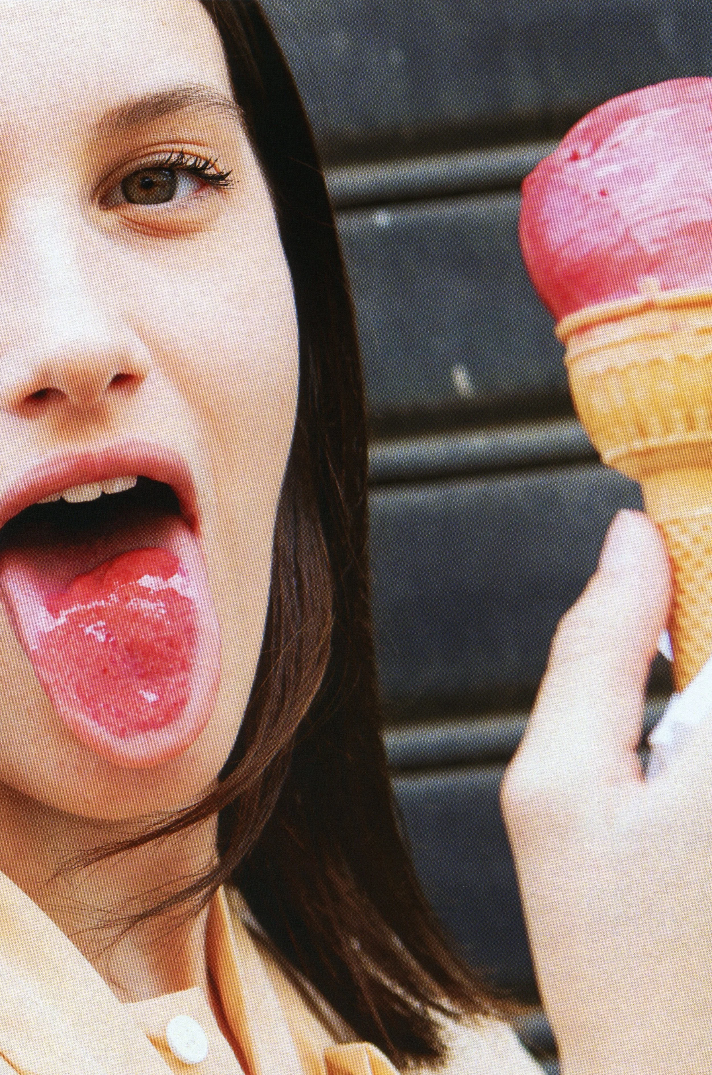 A woman with brown hair and light skin is licking a pink ice cream cone with her tongue out. The background is dark and blurred.