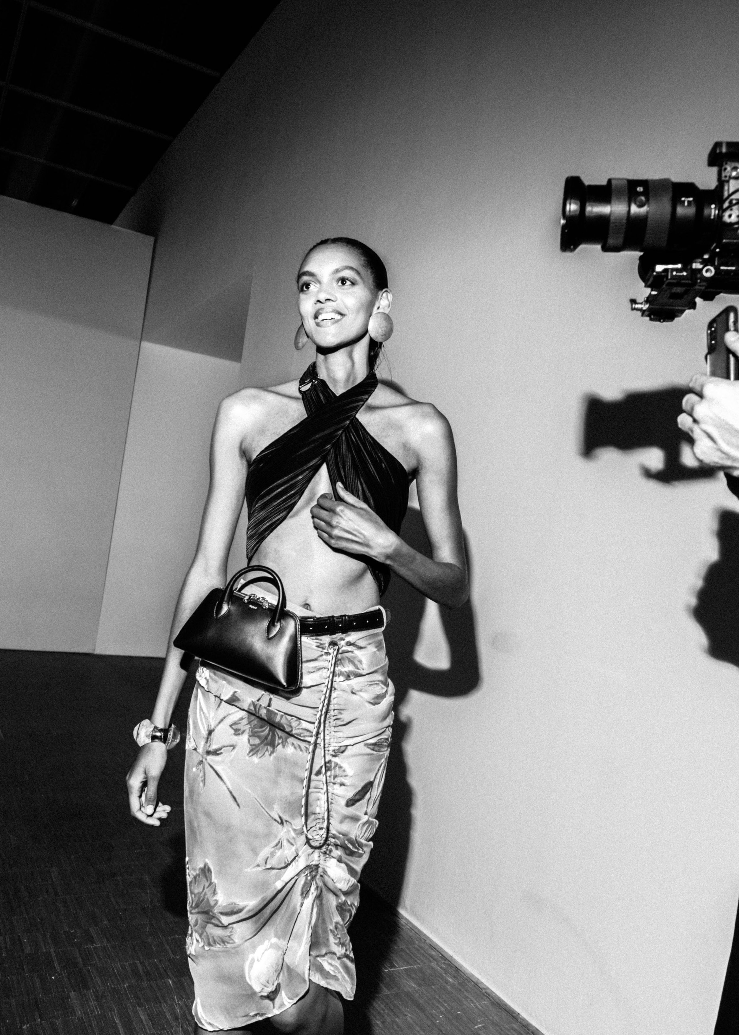 A woman with dark hair pulled back, wearing a cross-front black top, a floral satin skirt, large earrings, and a handbag, standing indoors in front of a plain wall with a photography camera pointed at her.