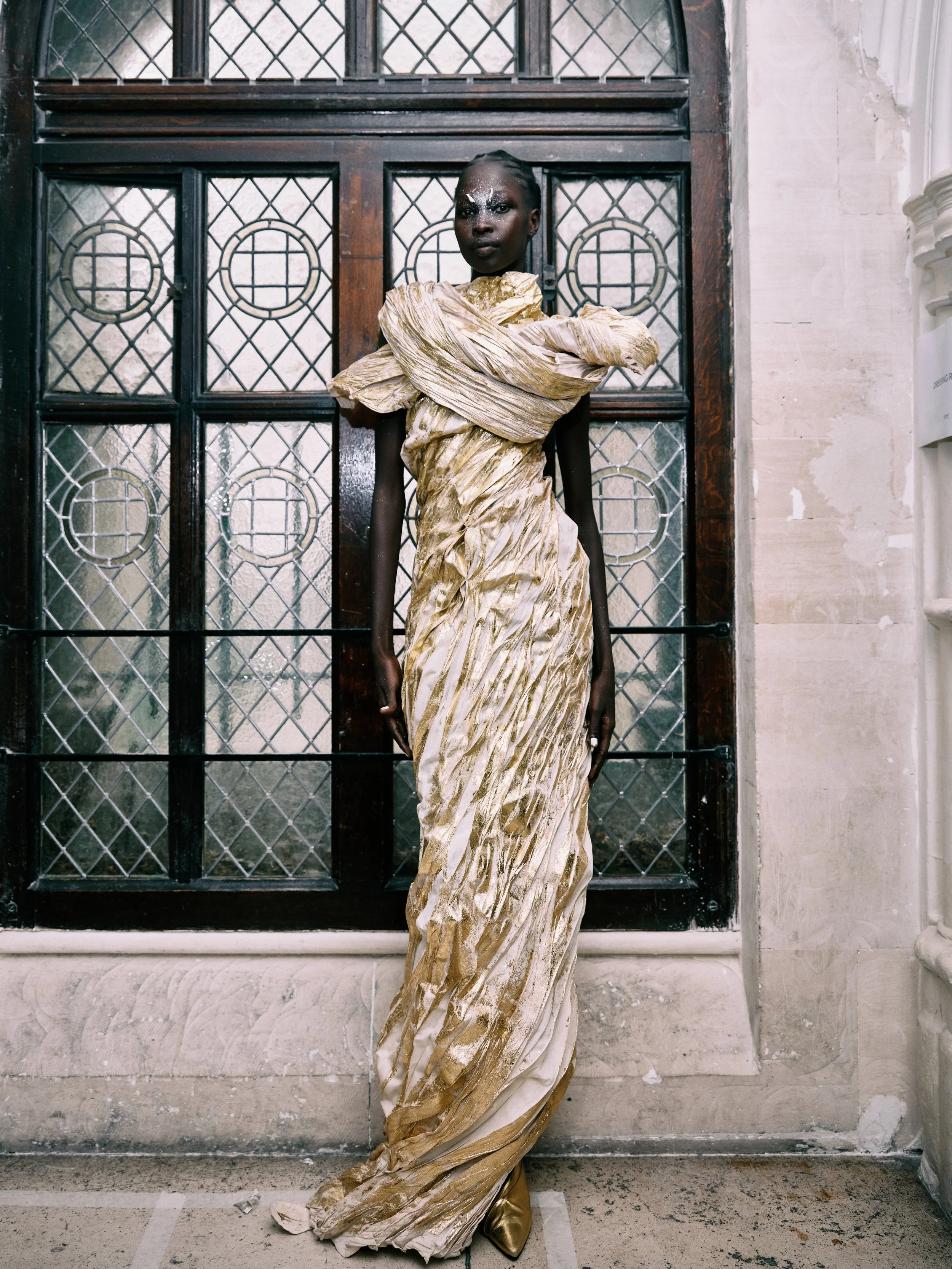 A person with dark skin and decorated face standing in front of a metal and glass door, wearing a gold and white fabric dress with a large draped design and metallic shoes.
