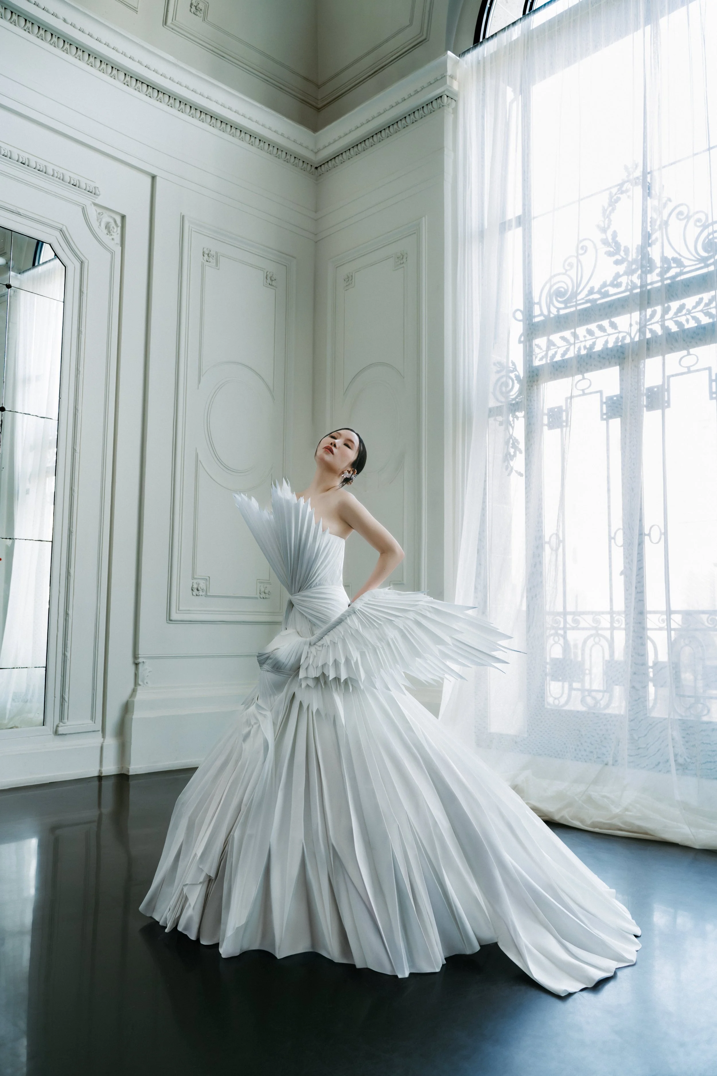 A woman in a white, pleated, gothic-style gown standing in a room with ornate white walls and large windows with sheer curtains, on a dark hardwood floor.