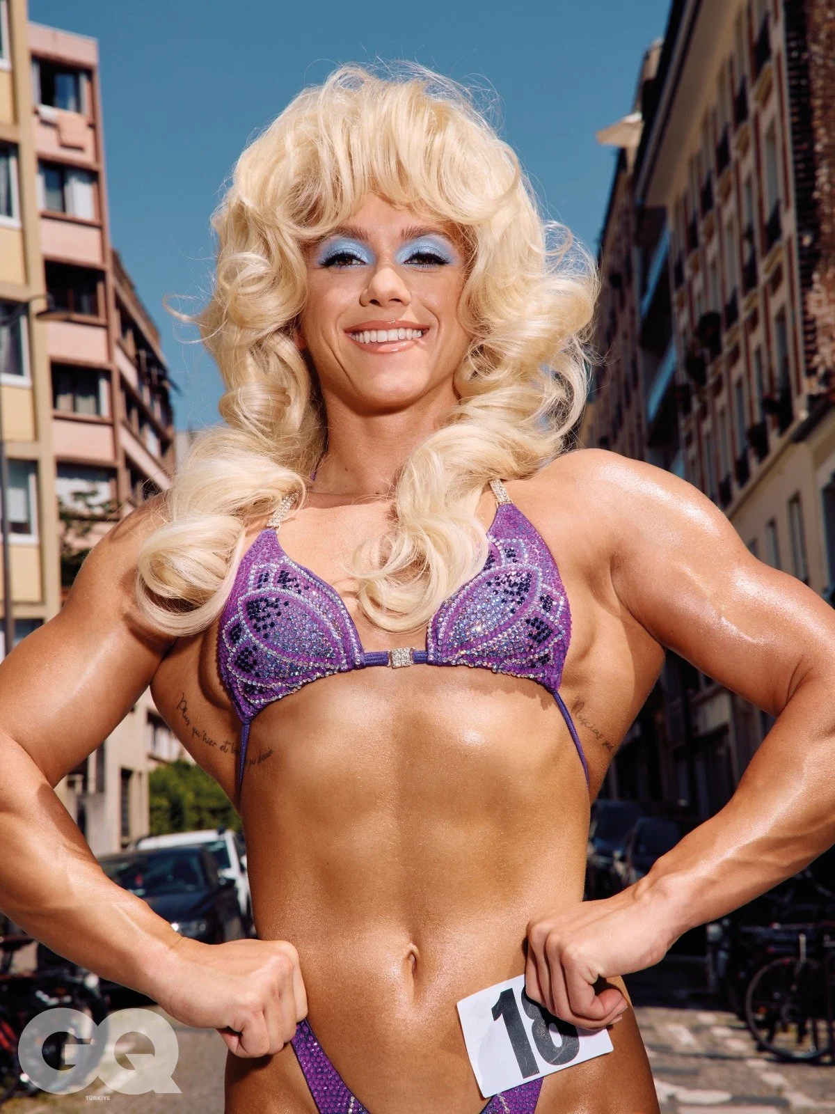 A muscular woman with long, curly blonde hair is smiling outdoors in a delta yellow and brown bikini top with purple rhinestone embellishments. She has a competition number on her waist and tattooed arms.