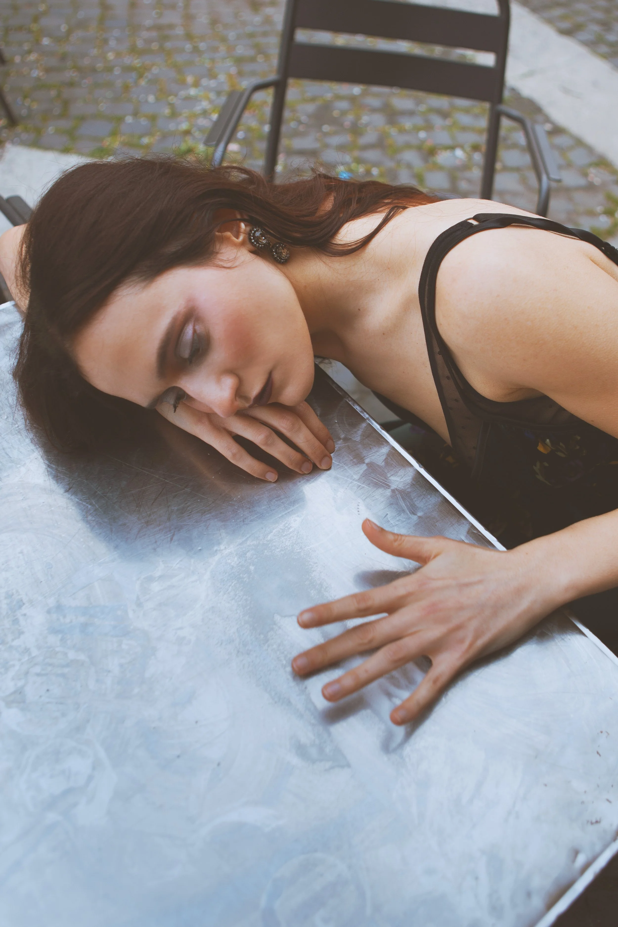 A woman with dark hair and earrings sleeping on a table outdoors, resting her head on her arm, with bare shoulders visible.