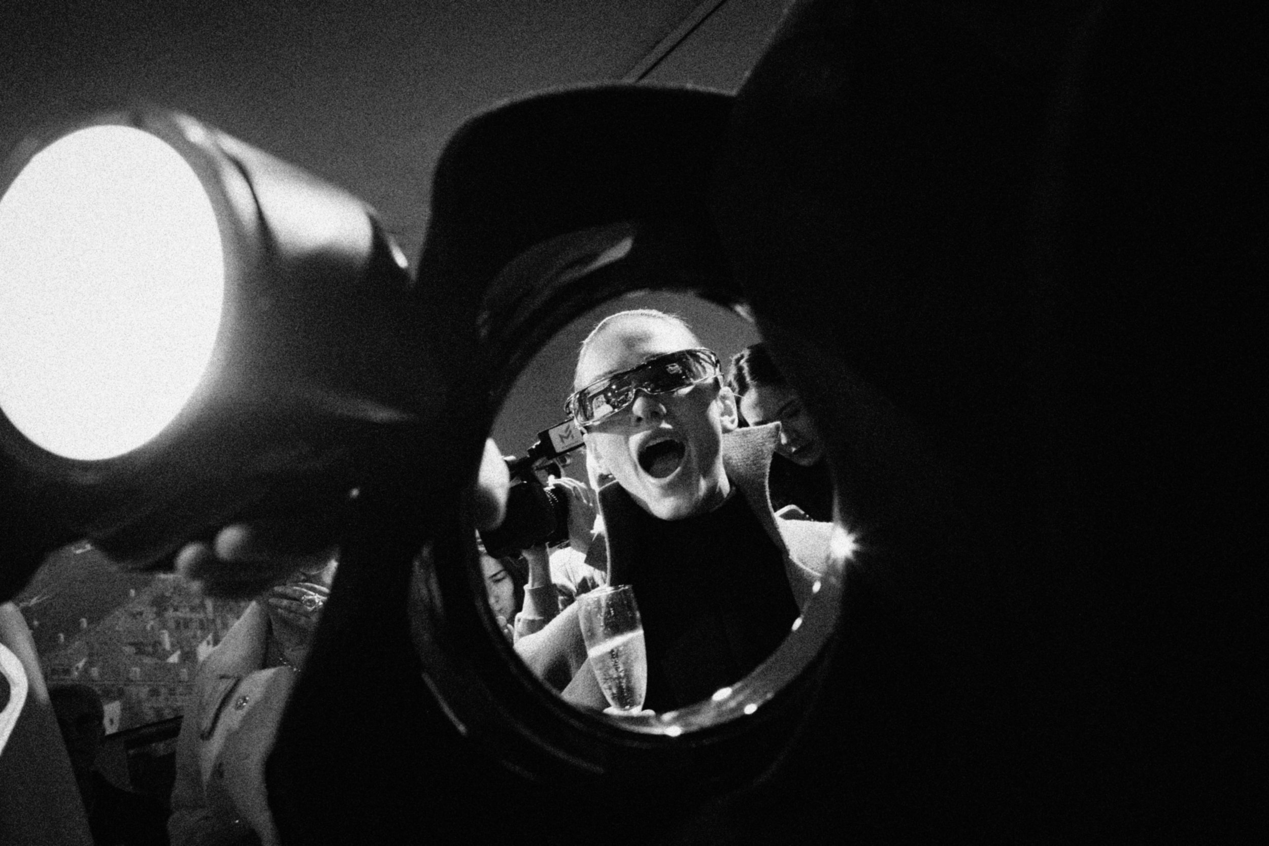 A person wearing sunglasses and a jacket is holding a camera and taking a selfie, with a woman holding a glass of champagne smiling behind her.