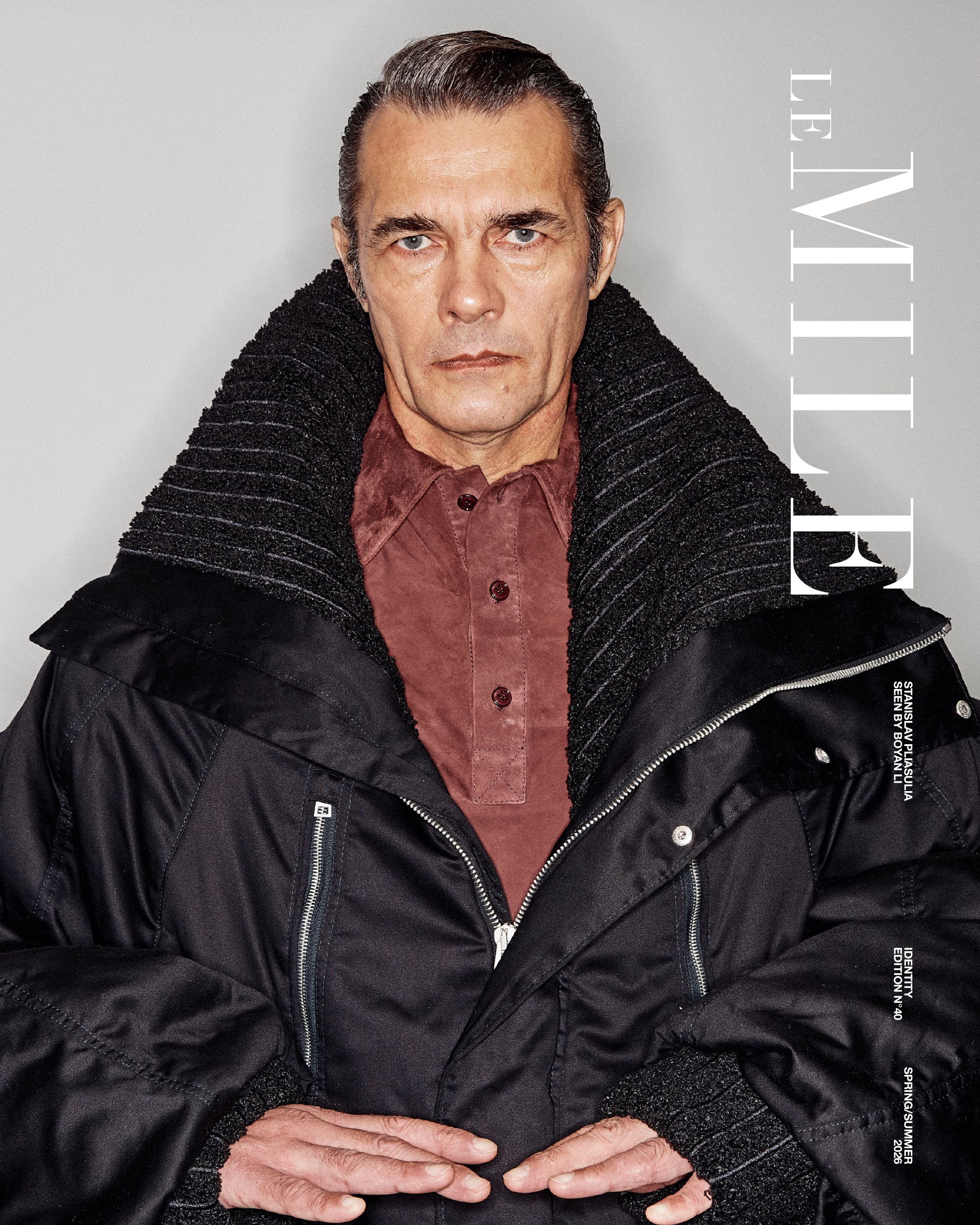 Close-up image of a middle-aged man with slicked-back hair, wearing a reddish-brown shirt and a thick black and ribbed jacket. He has piercing blue eyes and a serious expression, with his hands clasped in front of him. Vertical text on the right side reads "LEVI'S".