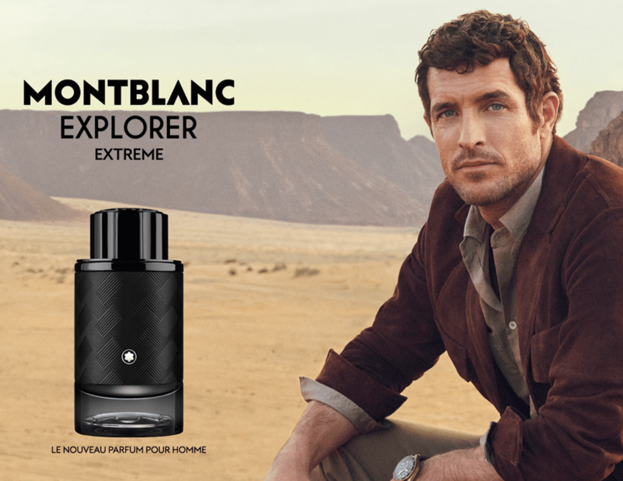 An advertisement for Montblanc Explorer Extreme men's fragrance features a man sitting outdoors in a desert landscape with mountains in the background. The man has curly brown hair, blue eyes, and is wearing a brown jacket over a light shirt. A black bottle of Montblanc Explorer Extreme perfume is displayed in the foreground, and the text indicates it is a new perfume for men.