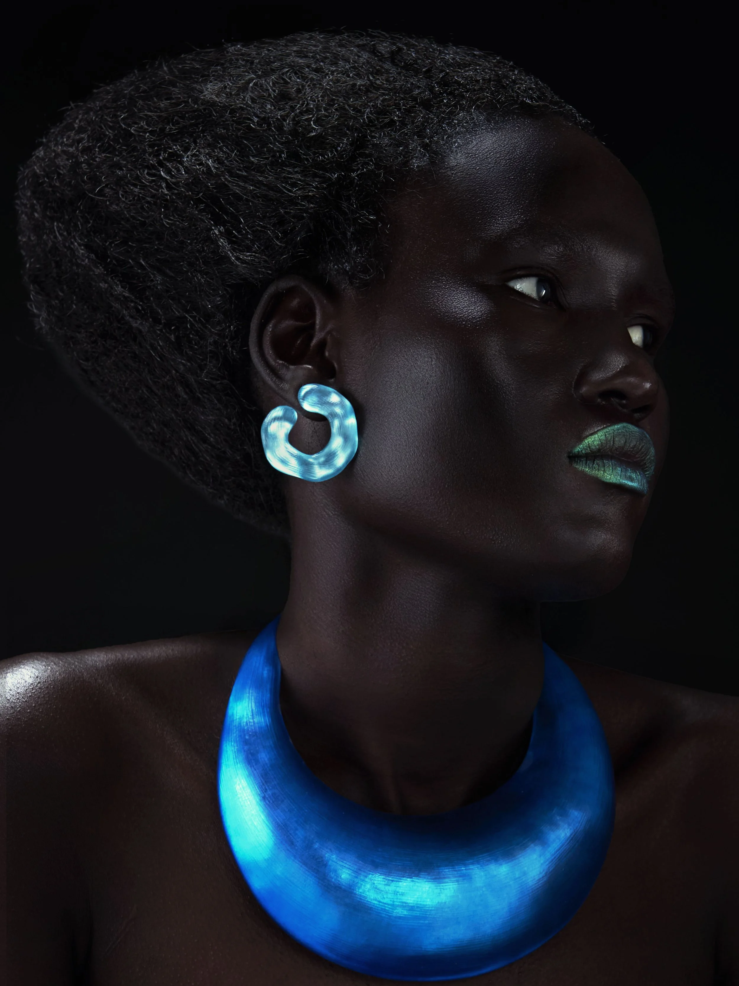 A close-up portrait of a Black woman with short, curly hair, wearing glowing blue earrings and a matching glowing blue necklace, against a black background.