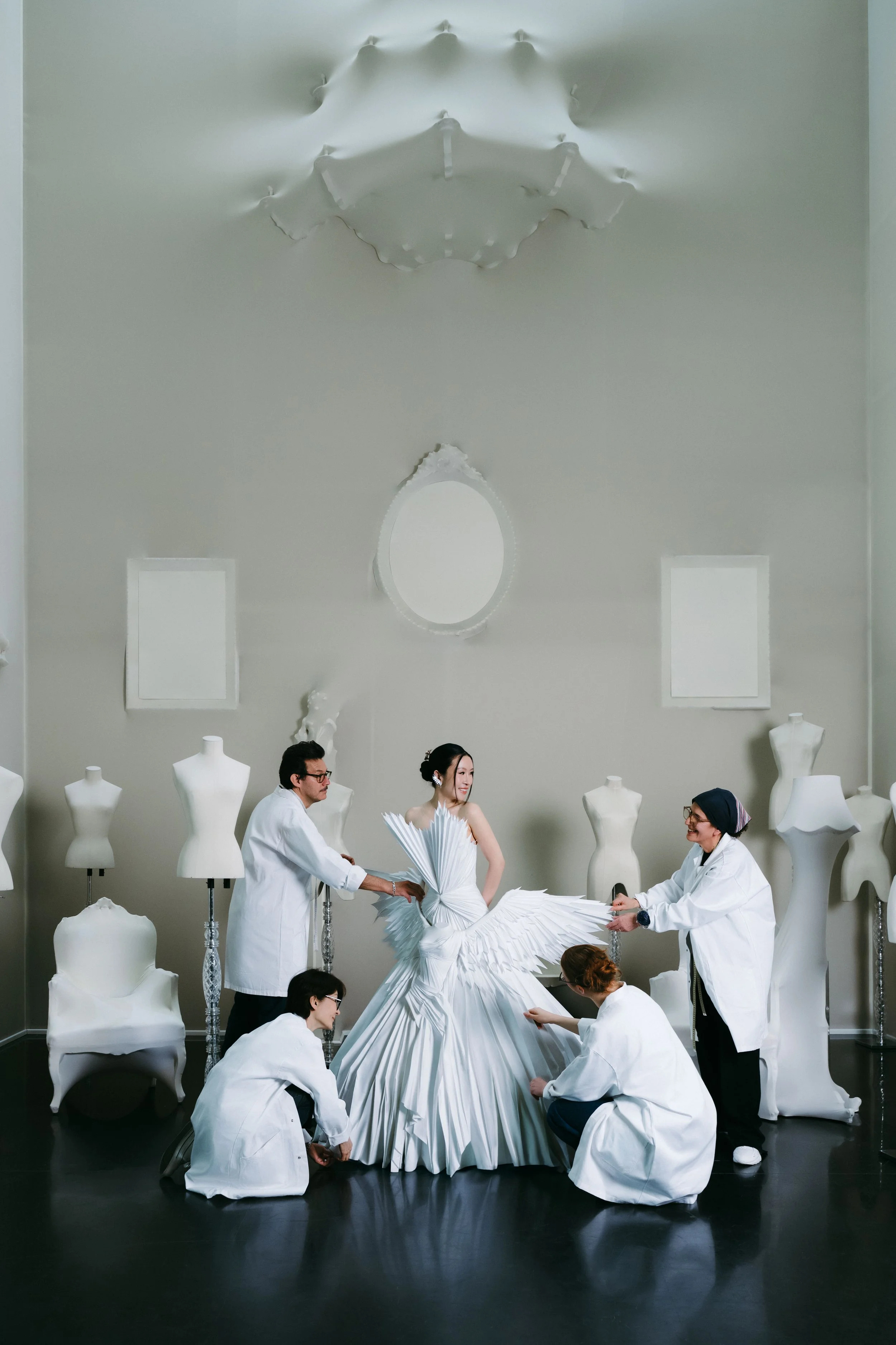 A performance scene with a woman in an elaborate white dress, surrounded by four people in white lab coats working on the dress in a room with white mannequins and a decorative ceiling fixture.