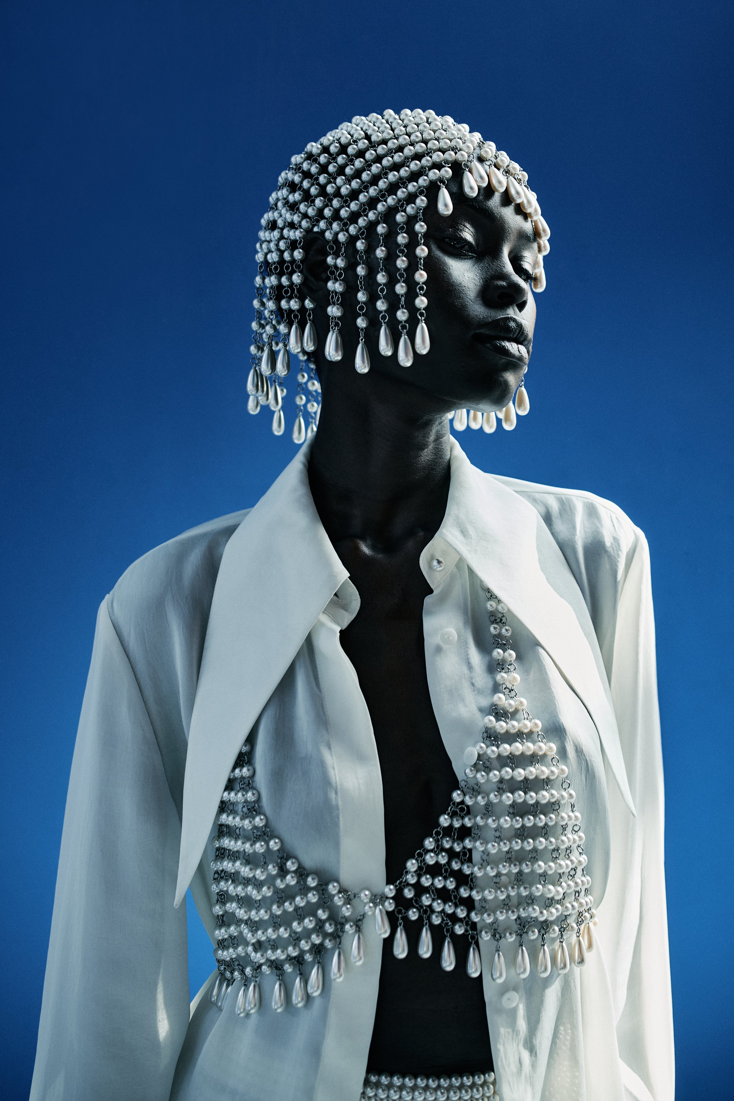 A person with dark skin and closed eyes, wearing a white shirt and a large pearl and silver bead headdress with teardrop-shaped pearl accents, against a solid blue background.