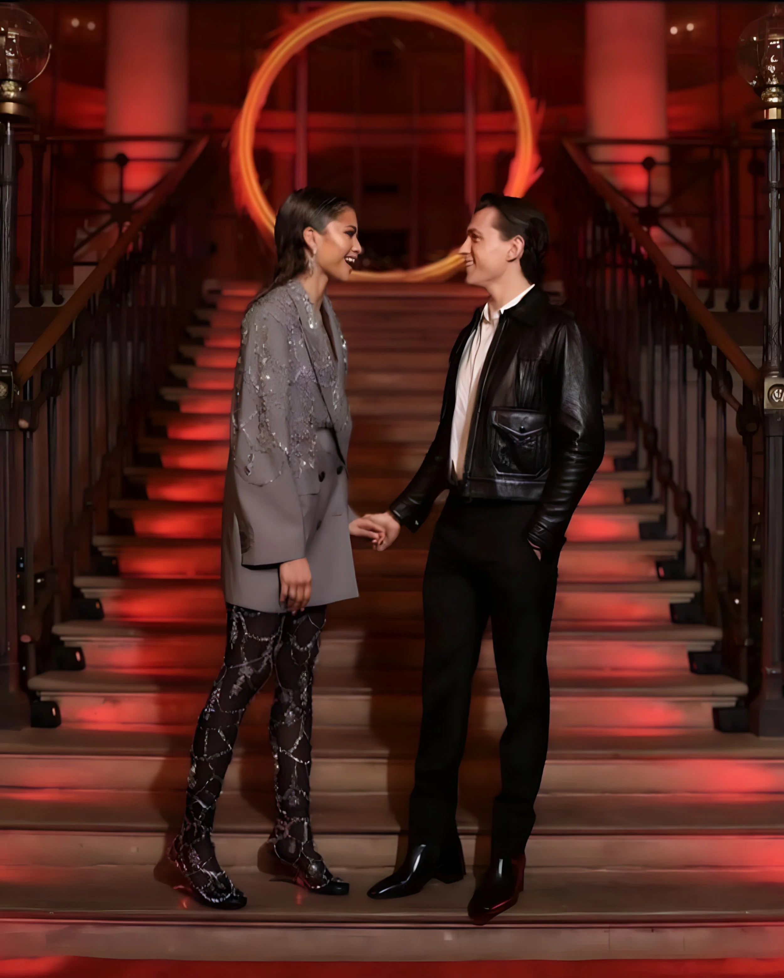 A man and woman smiling at each other while holding hands on a staircase with a glowing orange ring in the background.