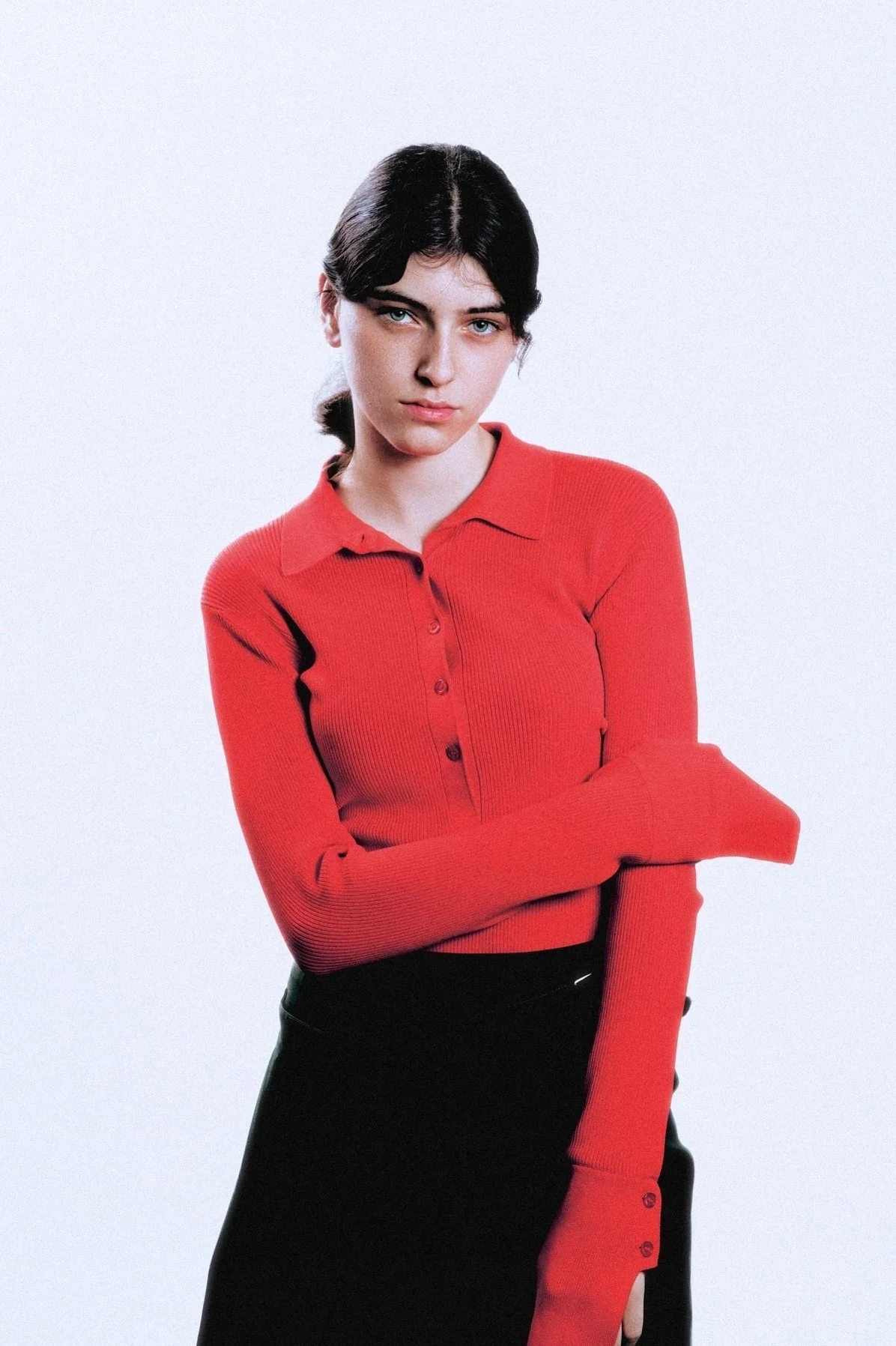 A young woman with dark hair wearing a red long-sleeve top and a black skirt stands against a plain light gray background.