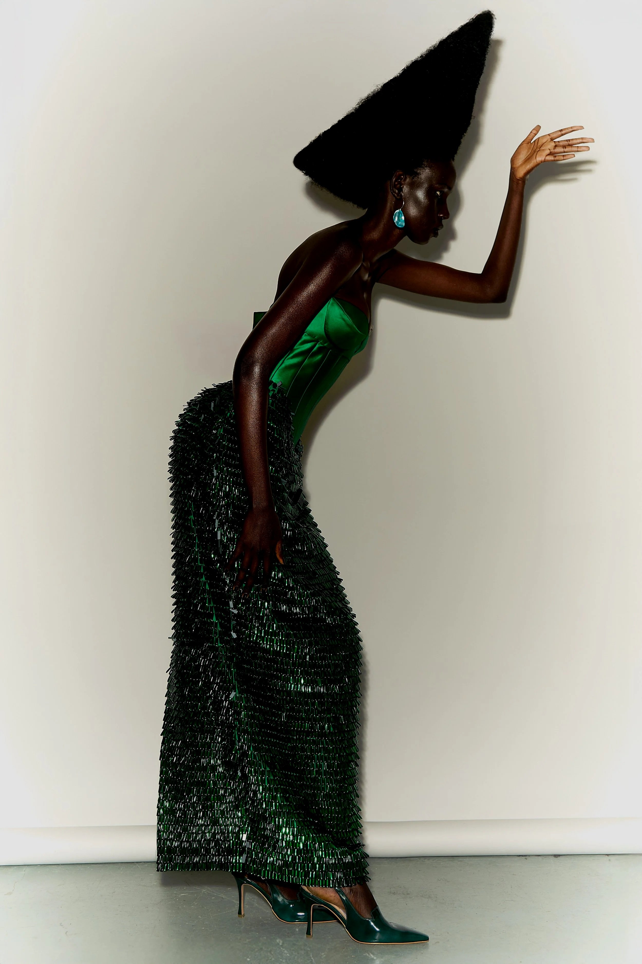 Fashion model with dark skin wearing a green strapless dress, beaded skirt, green high heels, and turquoise earrings, posing against a plain off-white wall.