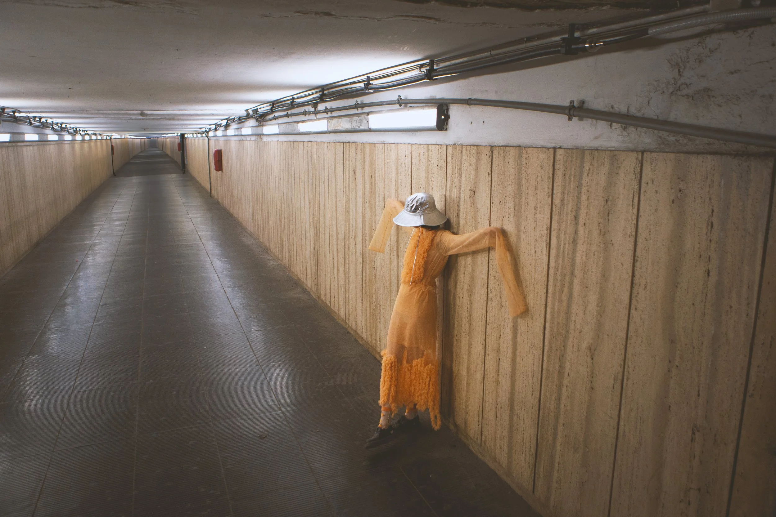 A long underground tunnel with tiled floor and wood-paneled walls, illuminated by rectangular lights on the ceiling. There is a scarecrow-like figure dressed in orange near the wall.