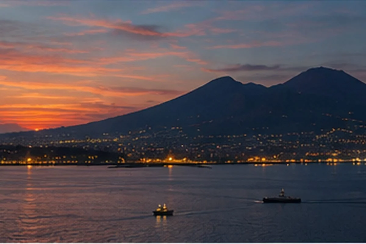 Mount Vesuvius at dusk across the Bay of Naples, with the mountain silhouetted against the evening sky and city lights glowing along the shoreline.