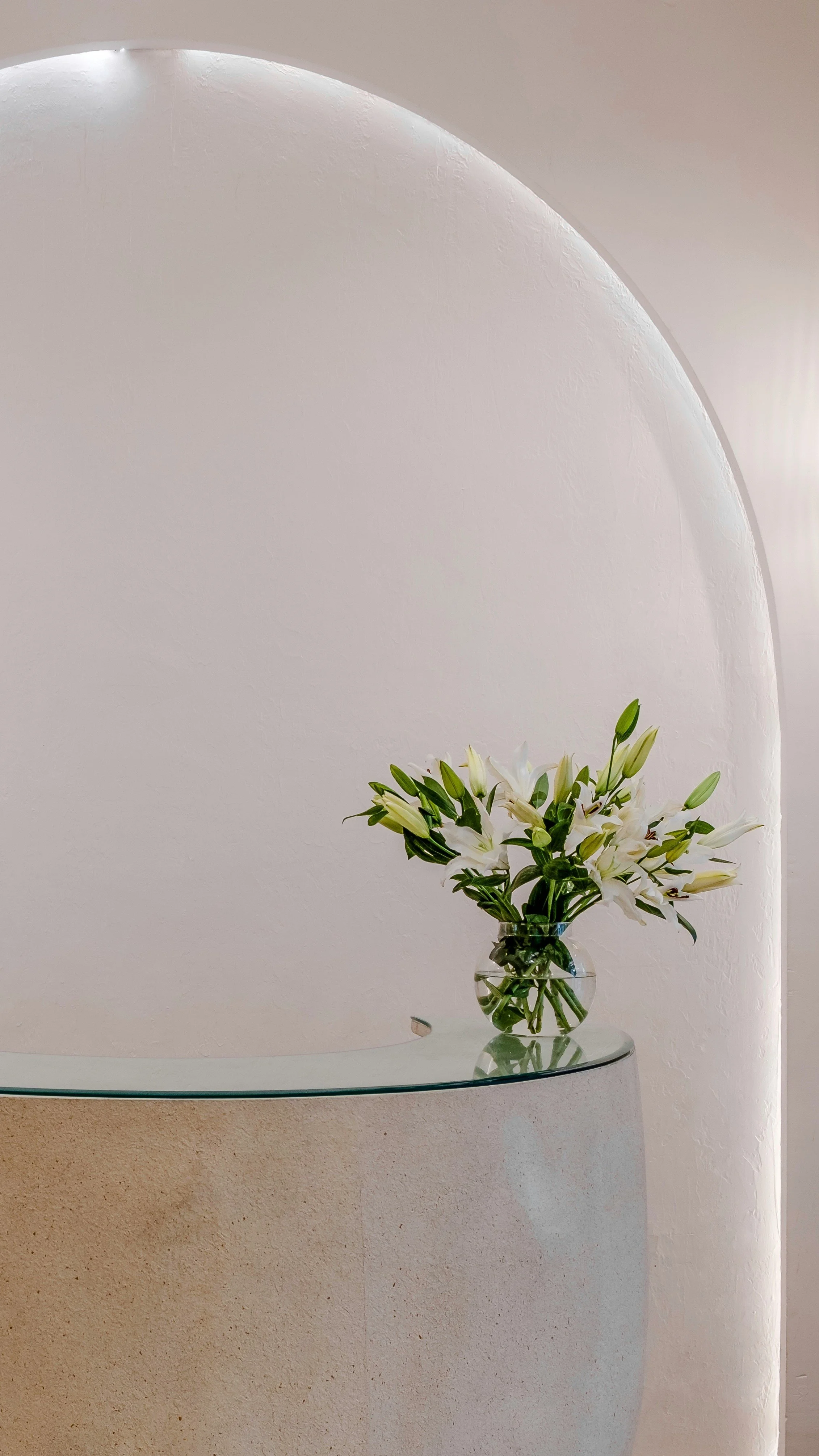 En vase med hvite liljer står på en glasskivebord mot en hvit vegg.