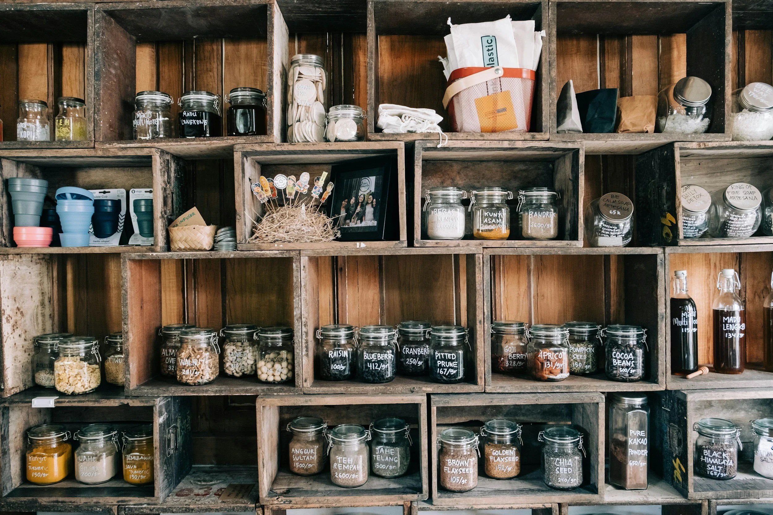 Wooden shelves holding glass jars with dry goods and spices, some labeled with handwritten tags, a framed photo, and a small straw basket.