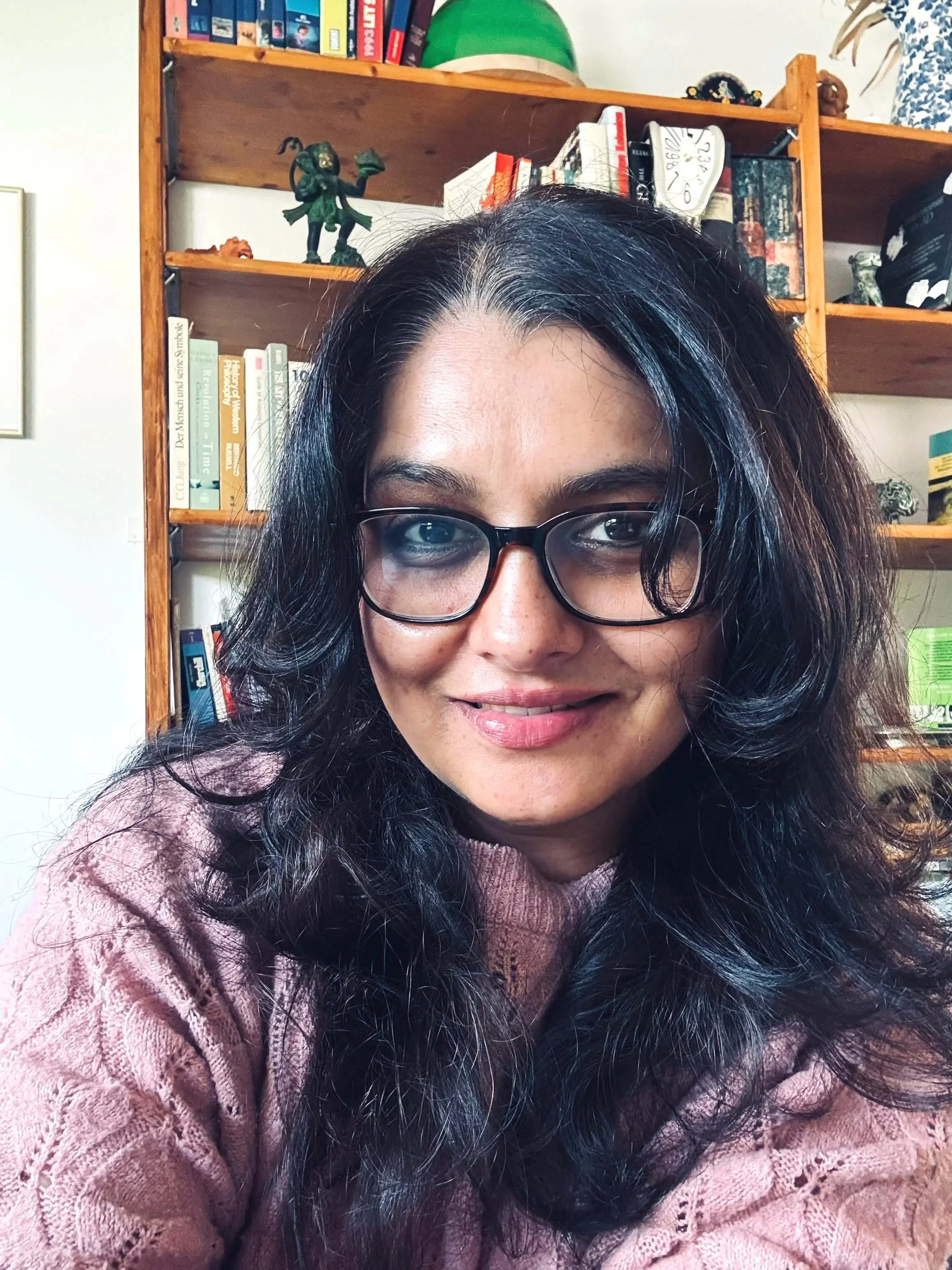 An Indian woman with brown skin and long hair, wearing glasses and sitting in front of a bookshelf