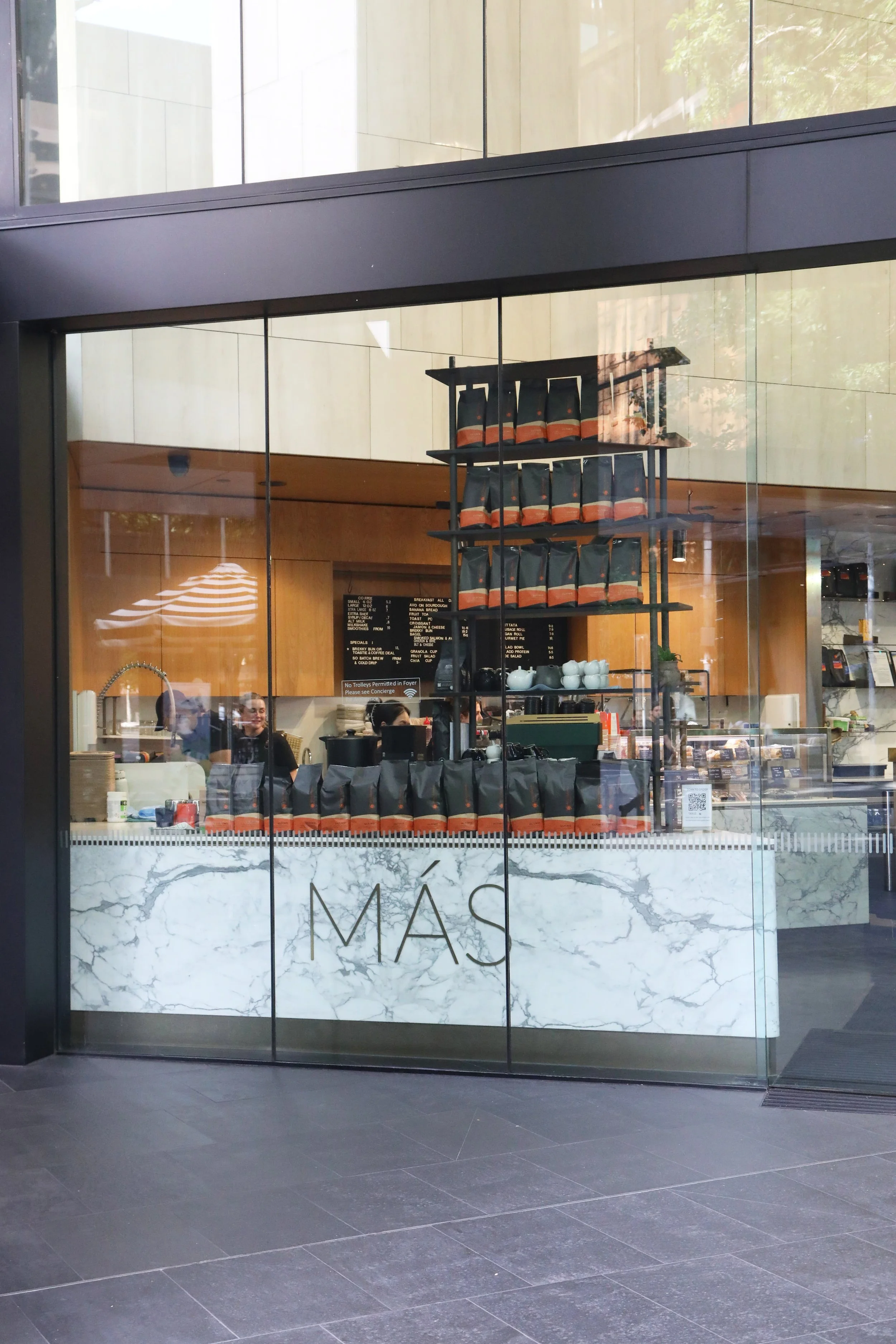 View of a modern cafe seen through a glass window, with shelves holding coffee bags and a menu visible inside.