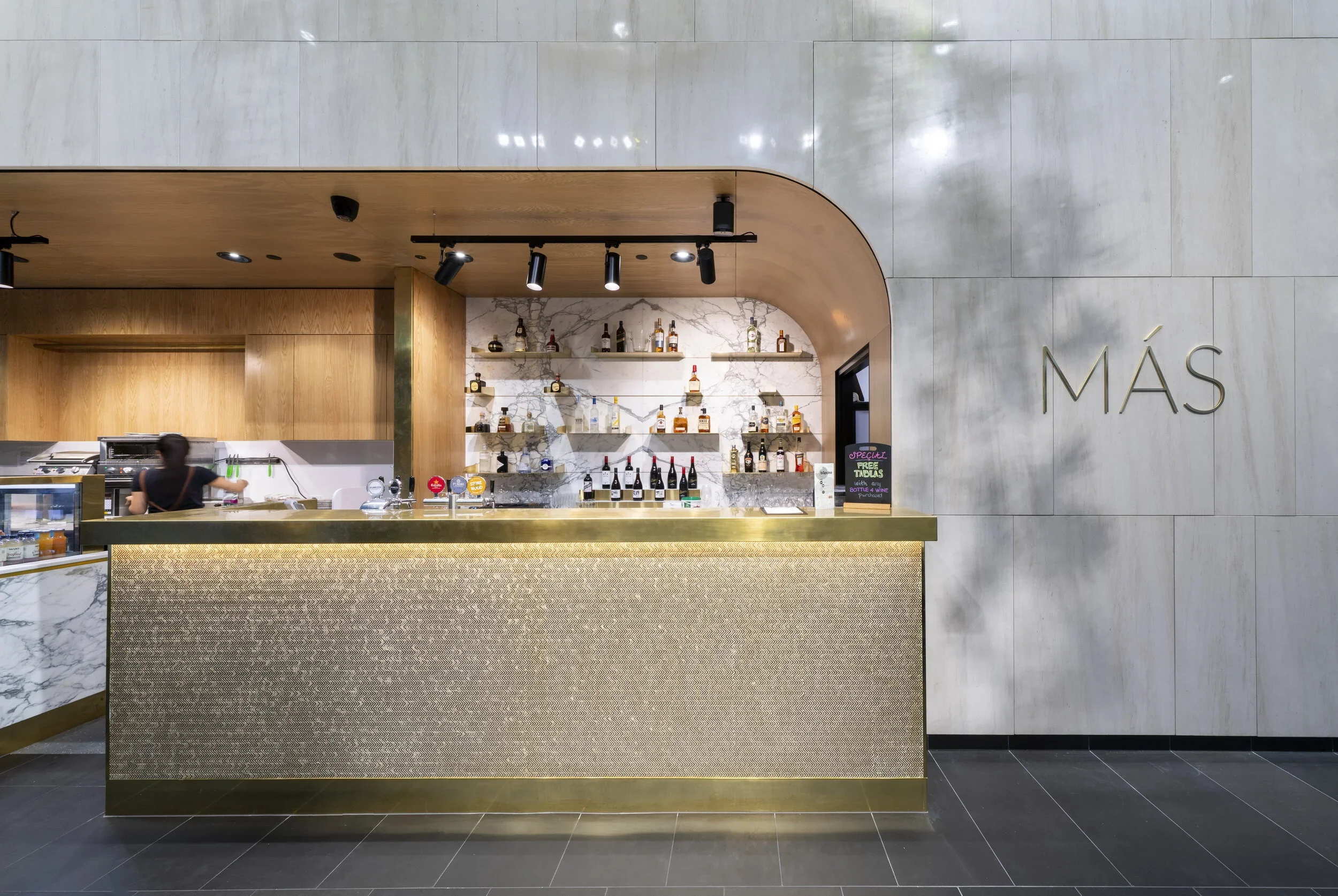 Modern bar area with a marble counter, wooden shelves with various liquor bottles, and a chef working behind the counter in a contemporary restaurant setting.