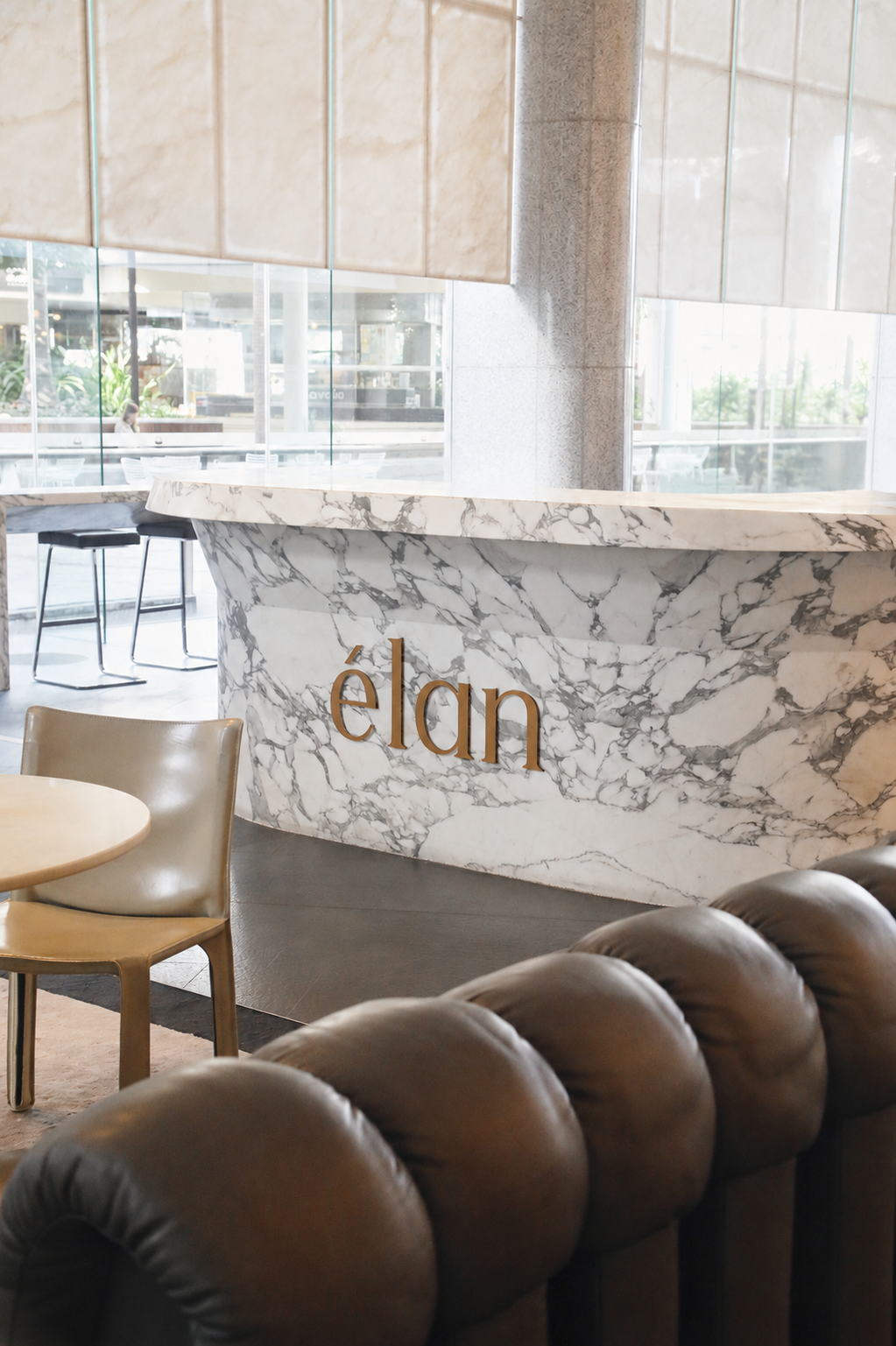 Marble reception desk with the word 'elán' in gold letters in a modern, stylish lounge area with beige and black chairs and large glass windows.