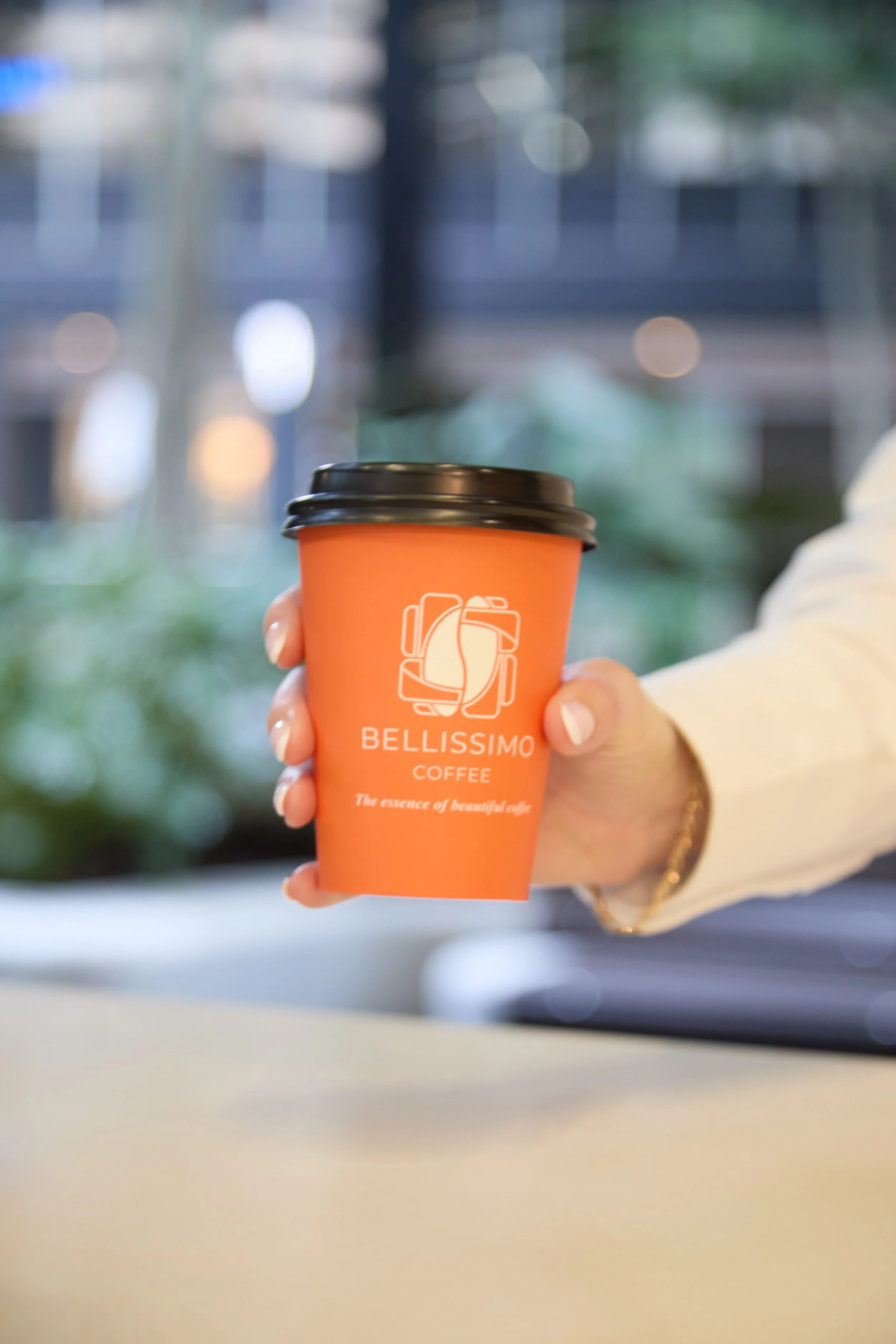 Person holding a bright orange coffee cup with the logo 'BELLISSIMO COFFEE' and the slogan 'The essence of beautiful coffee' on it. The person's hand is visible, with a gold bracelet on the wrist, and they are wearing a light-colored long sleeve.