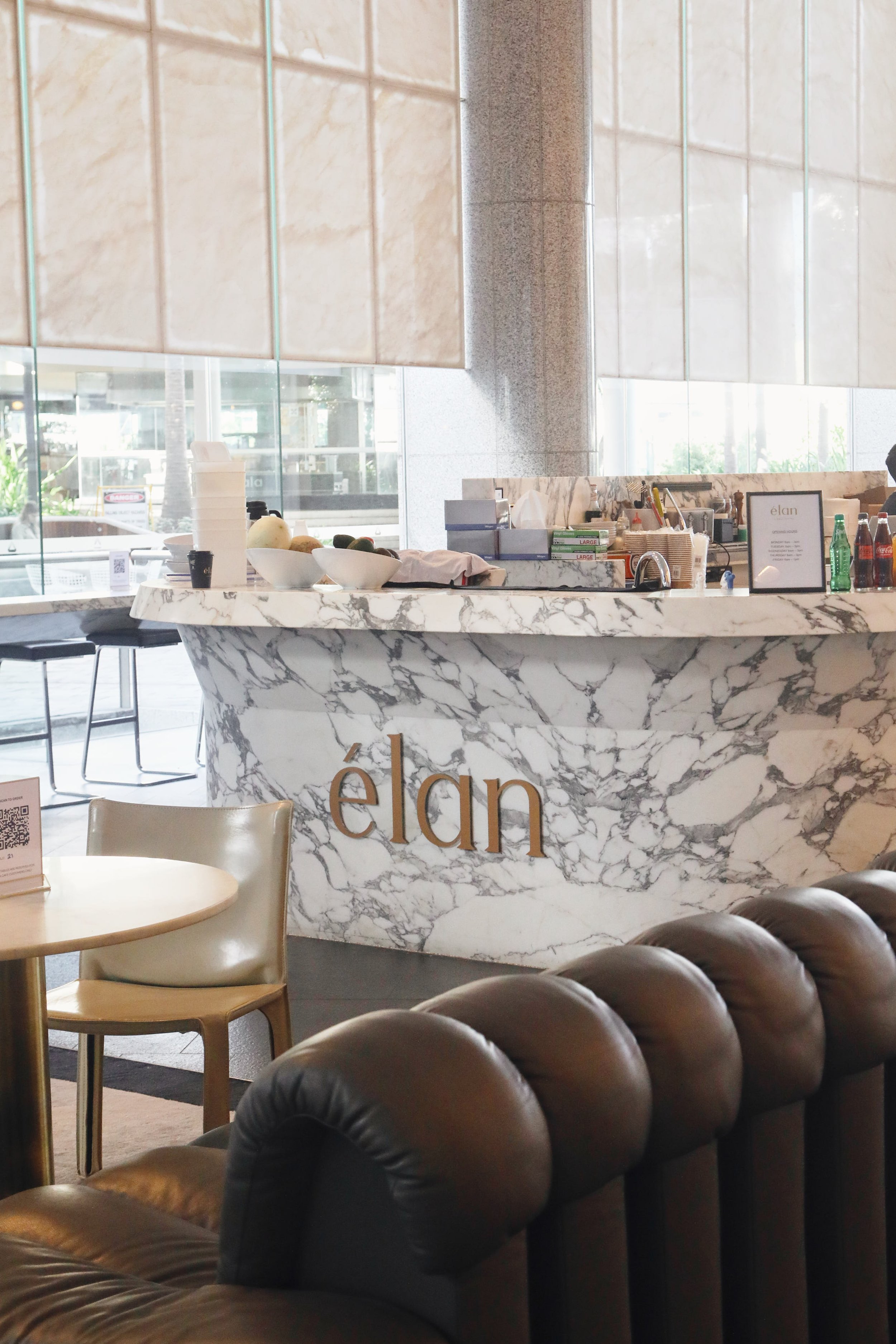 Interior of a modern cafe or restaurant with a marble counter, featuring stacks of paper cups, bowls of fruit, and beverage bottles, with plush black leather seating and a large window letting in natural light.