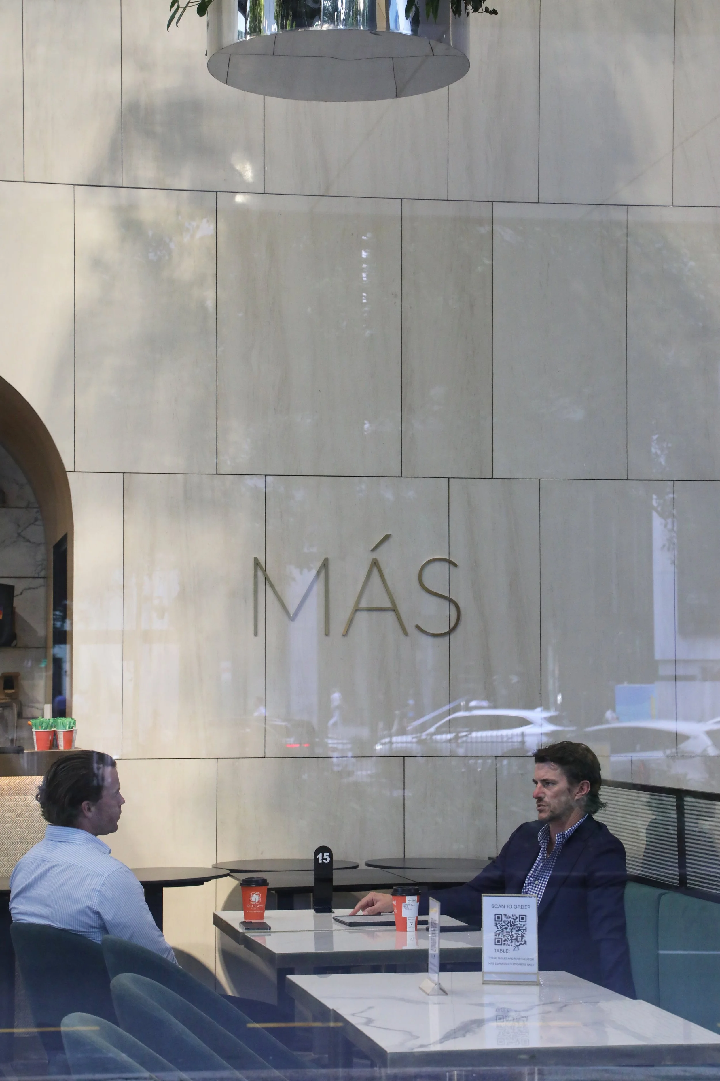 Two men sitting at a table in a modern cafe or restaurant with a large beige marble wall behind them. One man is wearing a white shirt, and the other is in a dark blazer. Drinks and a QR code sign for ordering are on the table. The environment looks contemporary and well-lit.