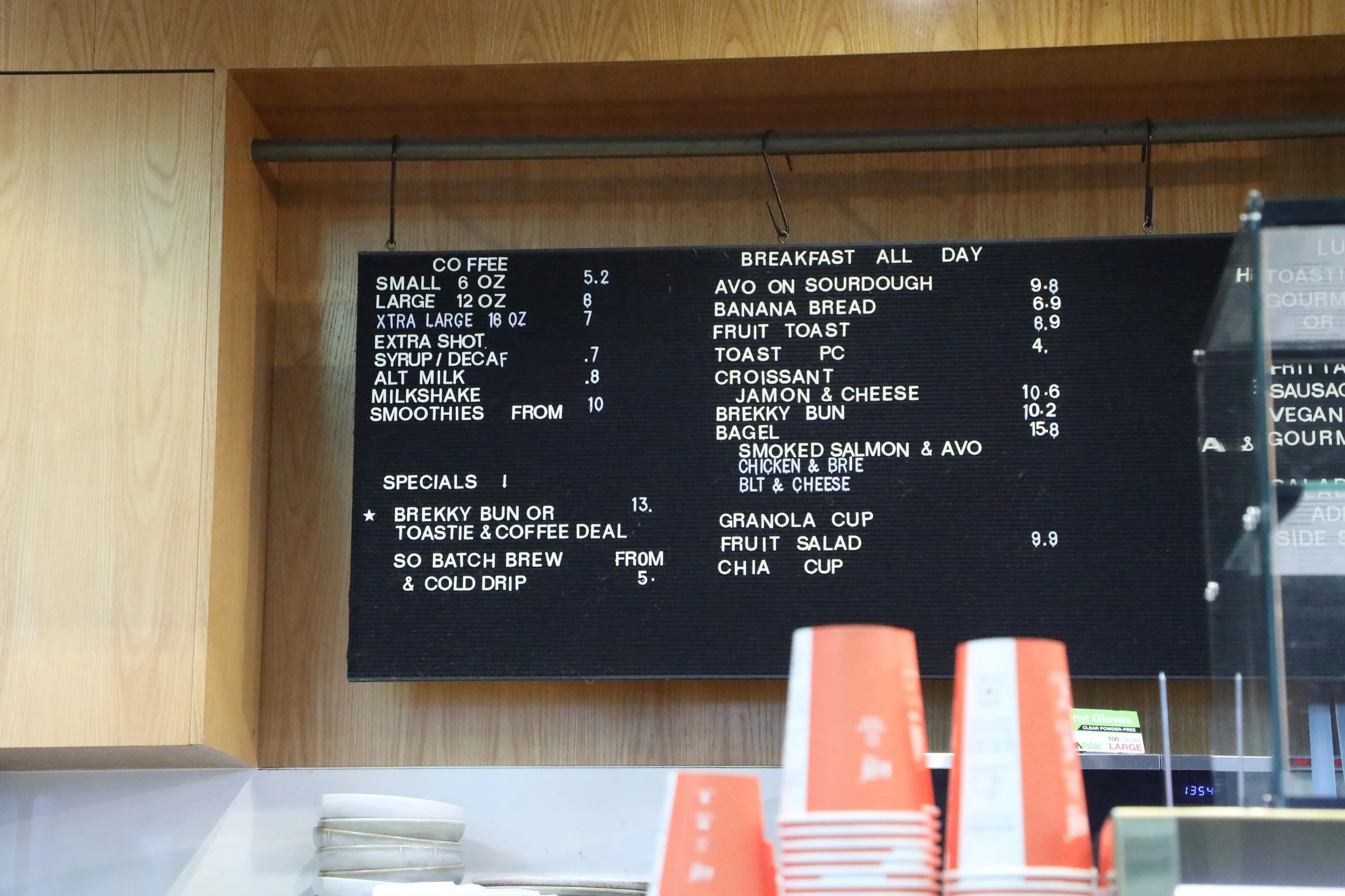 A black menu board listing beverages and breakfast items, with some orange and white product packaging partially visible in the foreground, mounted on a wooden wall.