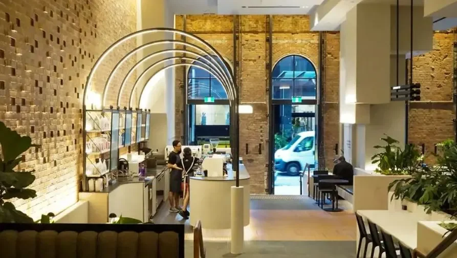 Interior of a modern cafe or bakery with brick walls, large arched windows, and a lit decorative arch structure above the counter. Staff and customers are present, with some seated and others ordering or preparing food.
