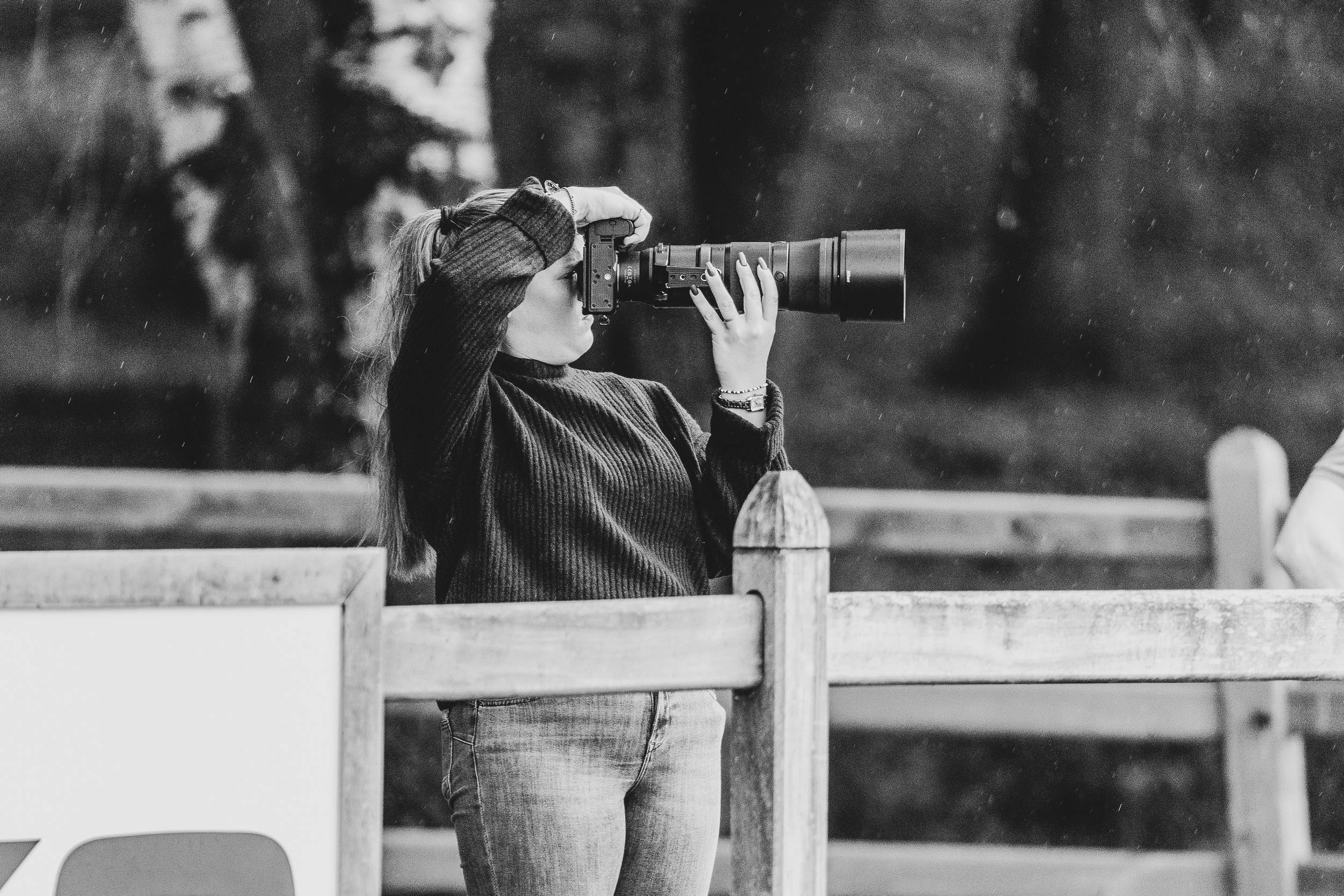 A woman wearing a sweater and jeans is holding a camera up to her face, taking a photograph outdoors in what appears to be a rainy environment.