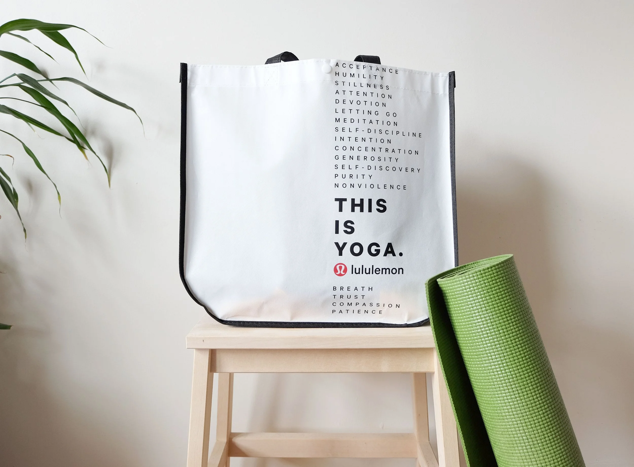 An interior scene with a white tote bag on a small wooden stool, a green yoga mat rolled up leaning against the stool, and part of a leafy green plant on the left. The tote bag has words related to yoga, mindfulness, and self-care printed on it, with some of the words visible including 'acceptance,' 'humility,' 'stillness,' 'attention,' 'devotion,' and 'yoga.'