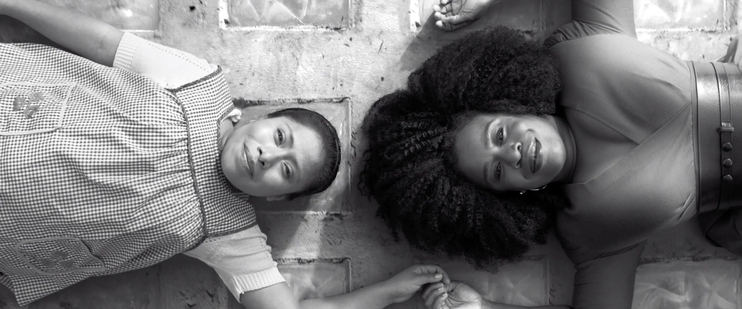 Two women lying on the ground holding hands, with one woman smiling and the other looking content, beside a brick wall.