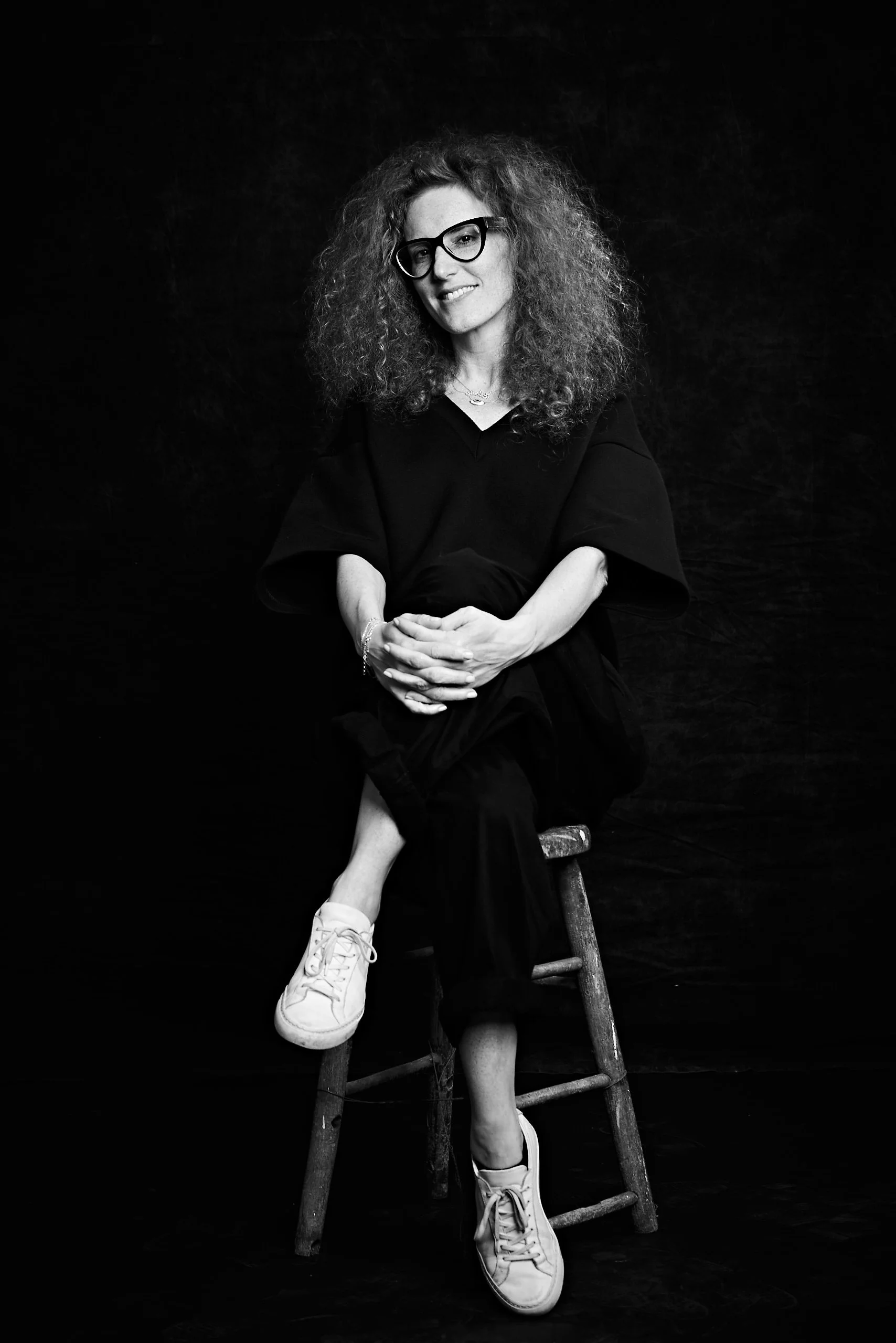 Black and white photo of a woman sitting on a wooden chair, wearing glasses, a black dress, and sneakers, with curly hair, posing against a dark background.