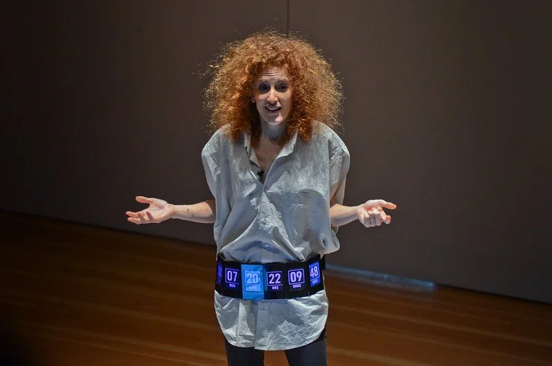 A woman with curly red hair wearing a white shirt and a digital belt displaying various numbers stands in a dimly lit room with wooden floors, gesturing with her hands.