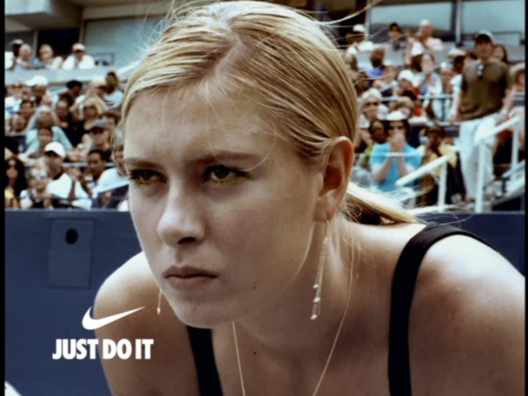 Close-up of a female athlete with blonde hair in a ponytail, wearing a black sports top, with a large crowd of spectators in the background at a sports event. Nike logo and slogan 'Just Do It' are visible.