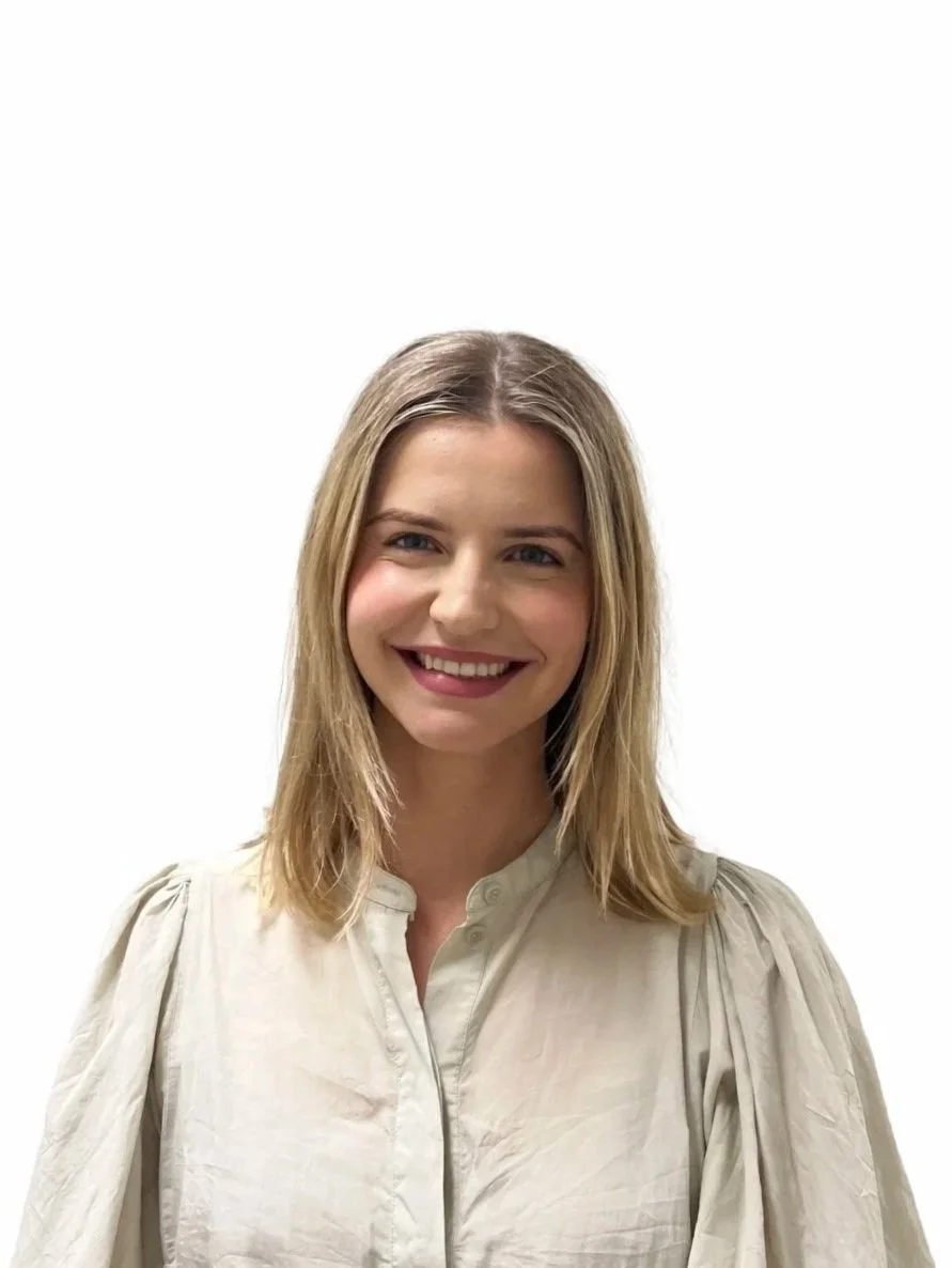 A woman with shoulder-length blonde hair smiling at the camera, wearing a light-colored blouse with puffy sleeves, against a plain white background.
