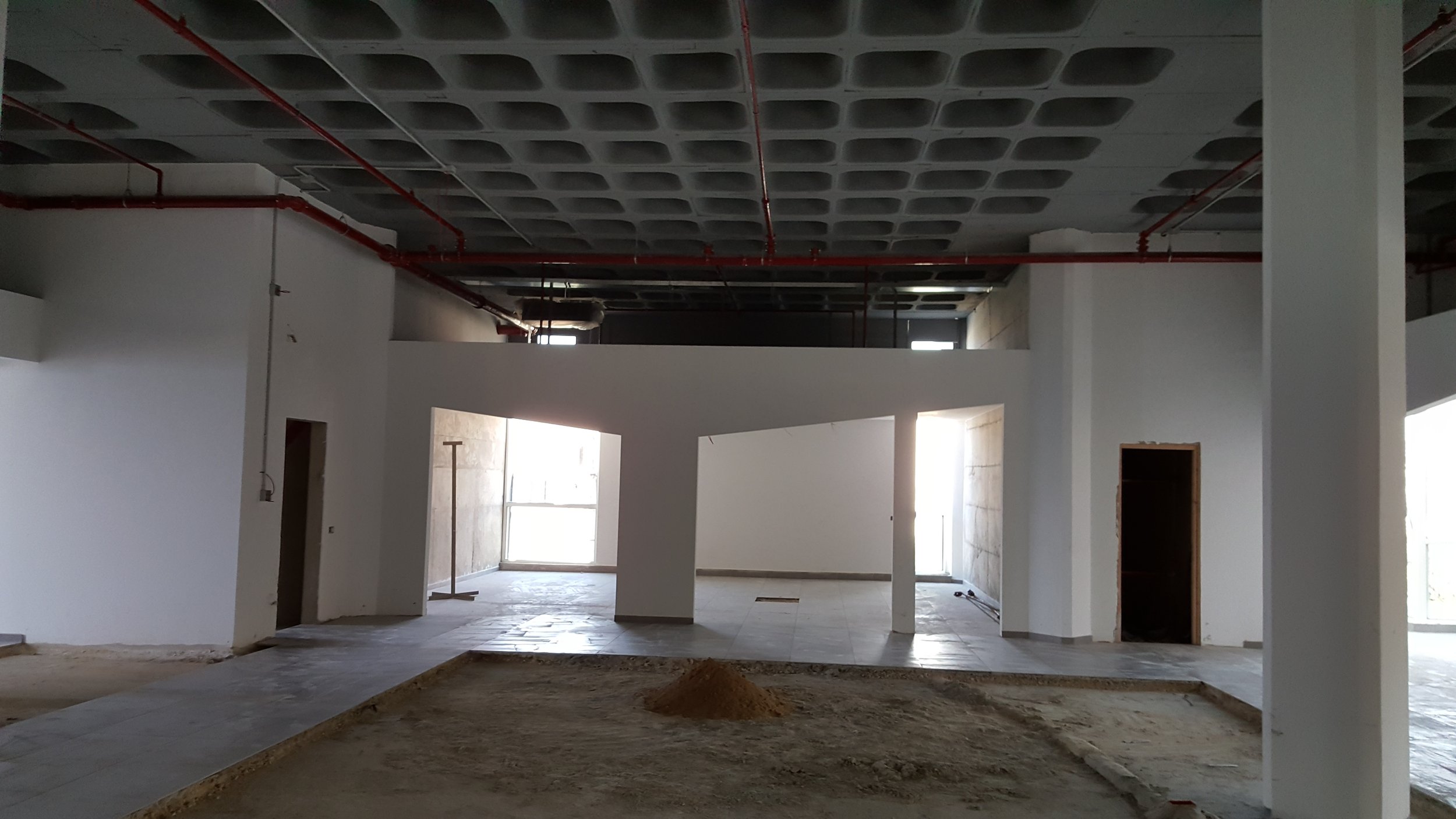 Interior of a building under construction with white walls, unfinished floors, exposed pipes on the ceiling, and large windows letting in natural light.