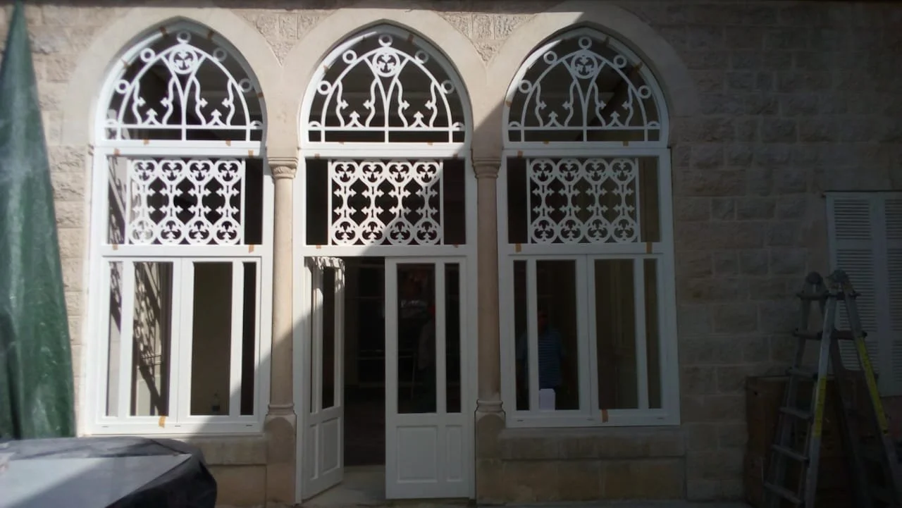 Three arched windows with decorative white ironwork on a stone wall. The middle window is open, revealing a dark interior. A ladder and some construction equipment are nearby.