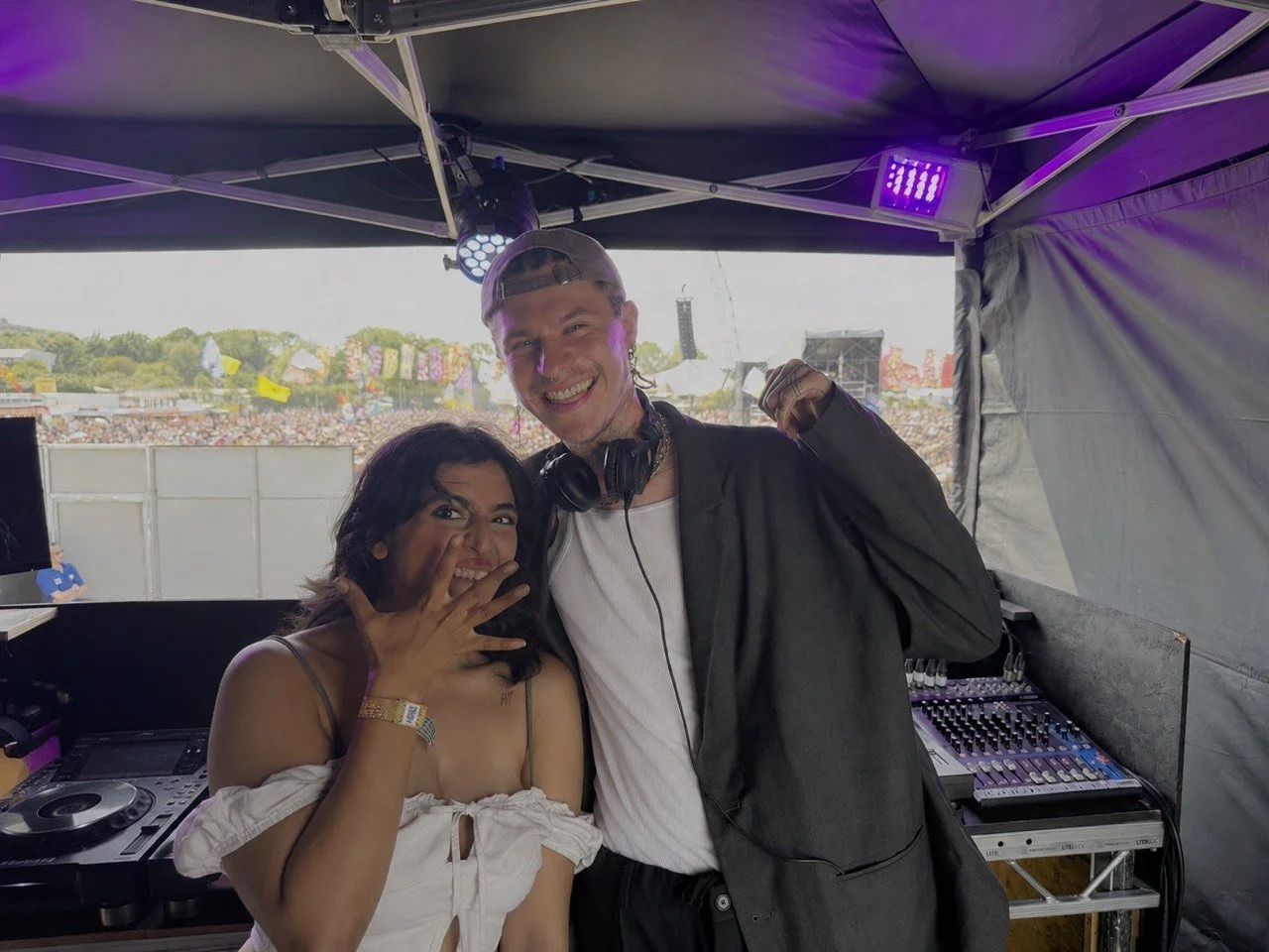 A woman and a man smiling and posing together at a music festival or outdoor event, with a crowd and fairground rides in the background, inside a DJ booth with equipment and purple lighting.