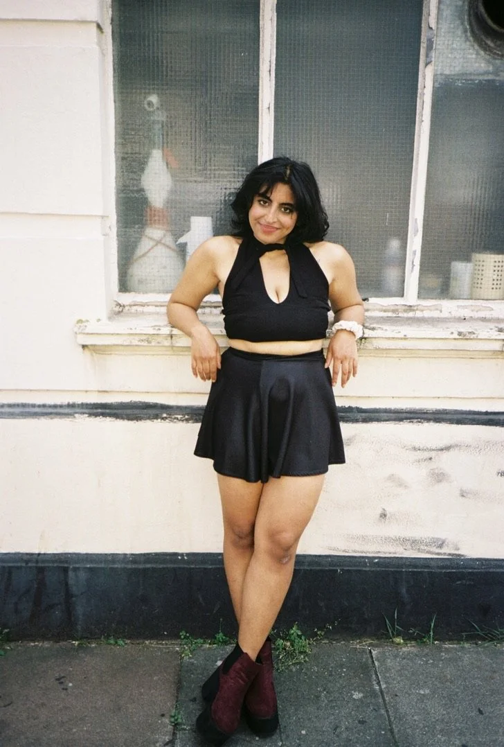 Young woman with shoulder-length black hair smiling, wearing a black halter top, black skirt, and burgundy ankle boots, standing in front of a window on a city sidewalk.