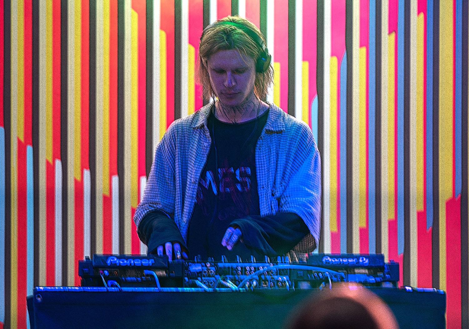 A DJ with shoulder-length blonde hair wearing headphones, a black graphic t-shirt under an open plaid shirt, mixes music at a vibrant event with colorful, striped background lighting.