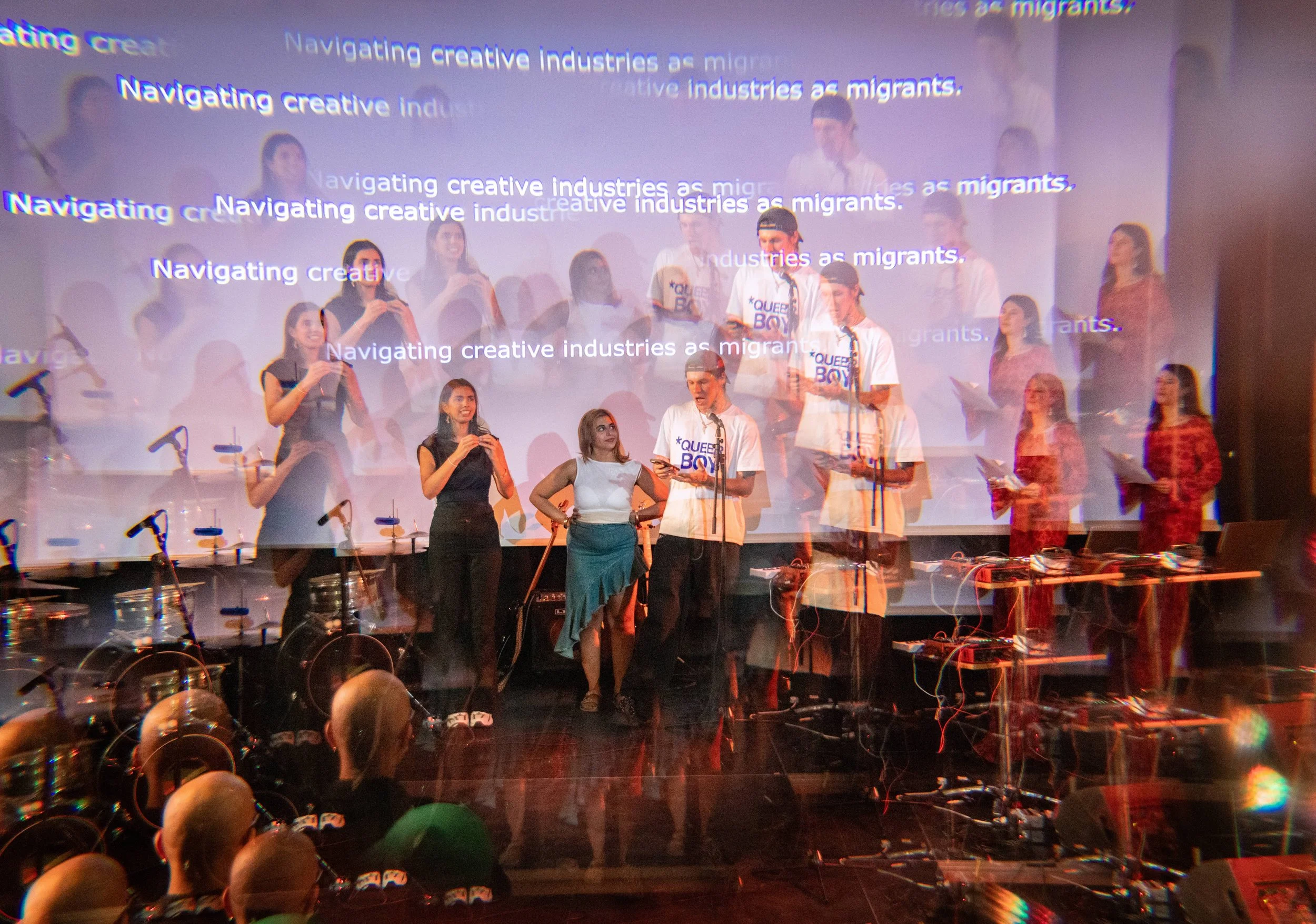 A group of people on stage, some holding papers, with a large screen behind them displaying multiple lines of text that say "Navigating creative industries as migrants." The individuals are standing in front of a microphone stand, and there are musical instruments like drums and electronic equipment visible at the bottom of the image.