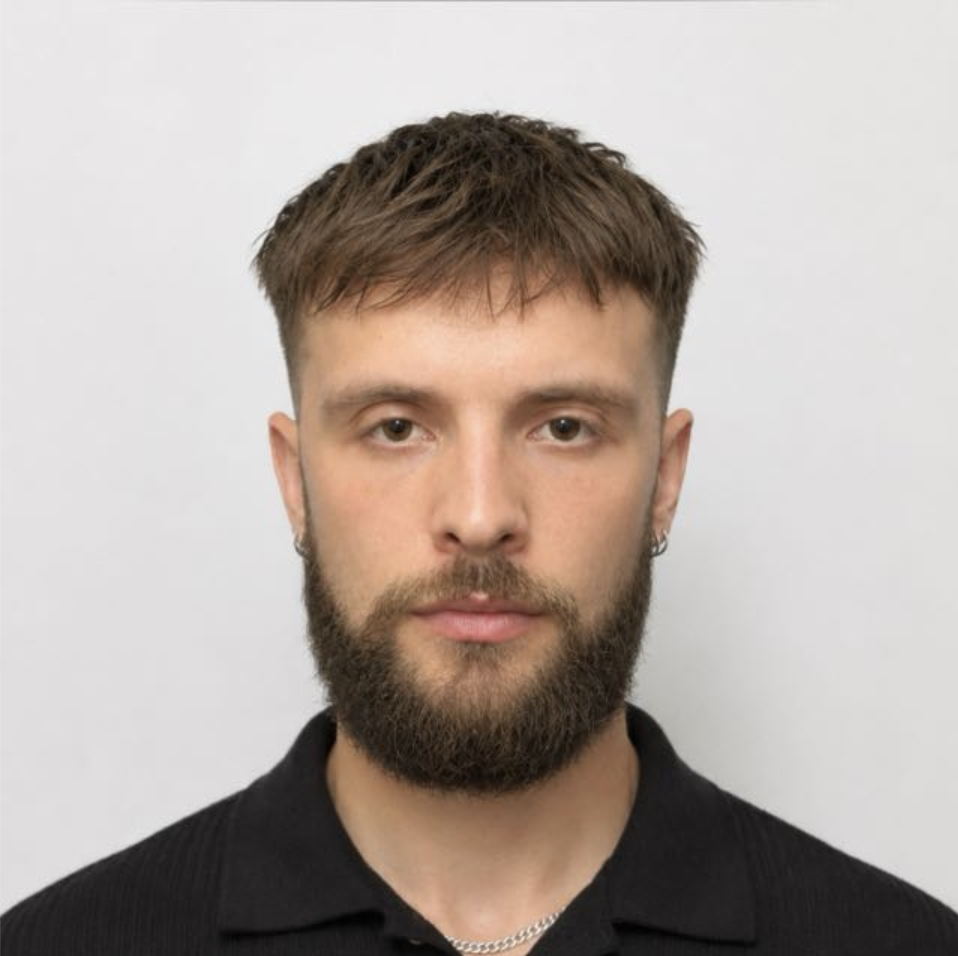 A portrait of a young man with light skin, short brown hair, a full beard, wearing a black shirt and a chain necklace against a plain white background.