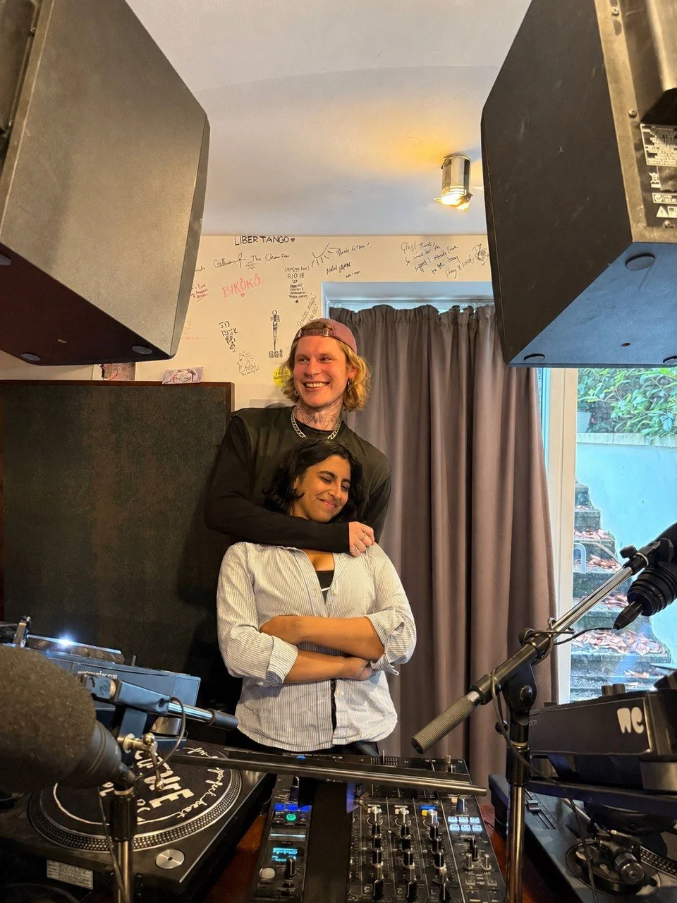 A man and a woman posing together indoors in front of DJ equipment, with a wall covered in handwritten notes and graffiti behind them.